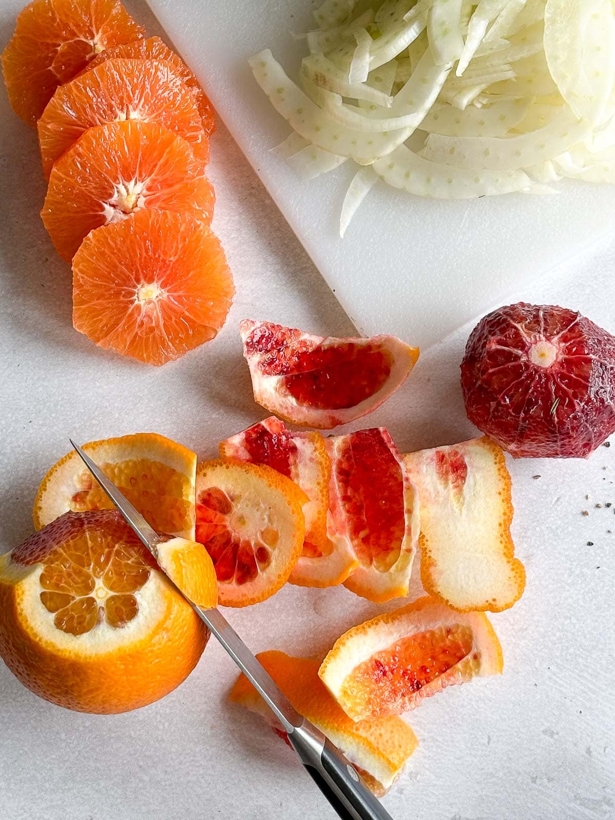 knife sliced peel off oranges next to orange sliced and peeled blood orange and board with pile of sliced fennel. 
