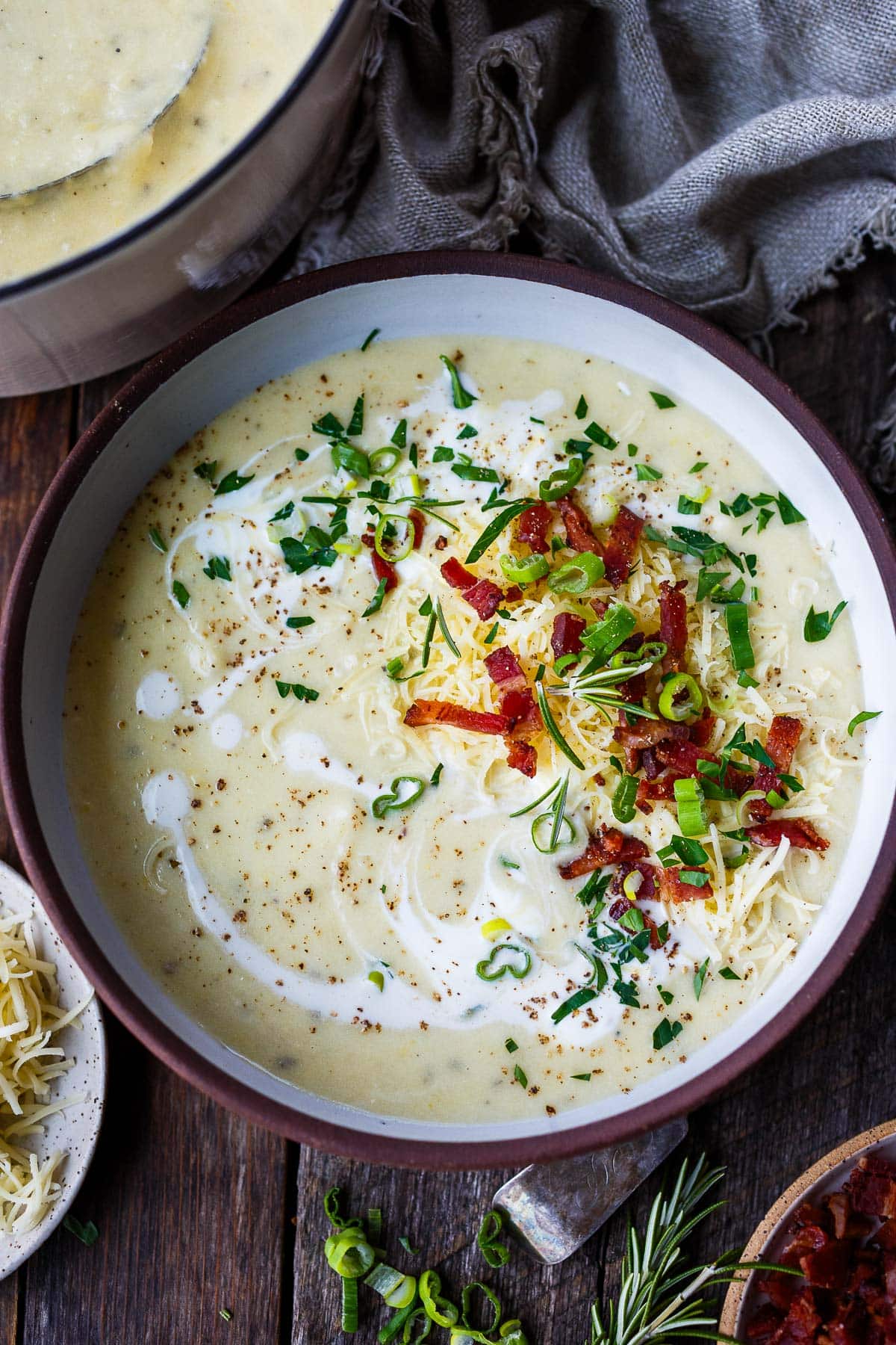 bowl of potato soup with swirl of cream, garnished with chopped chives, green onions, and rosemary, crispy bacon, and grated cheddar. 