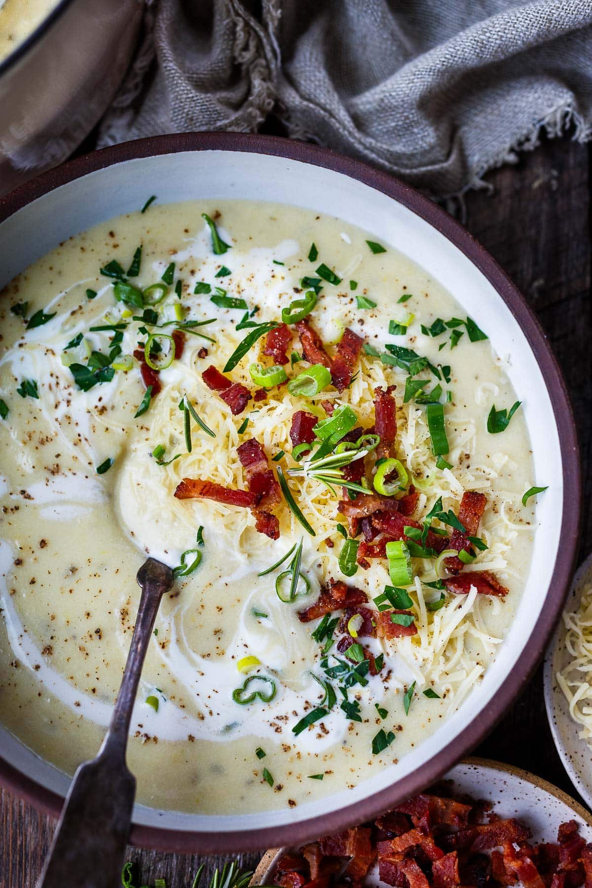 bowl of potato soup with spoon, garnished with bacon, green onions, chives, grated cheddar, and a swirl of cream.