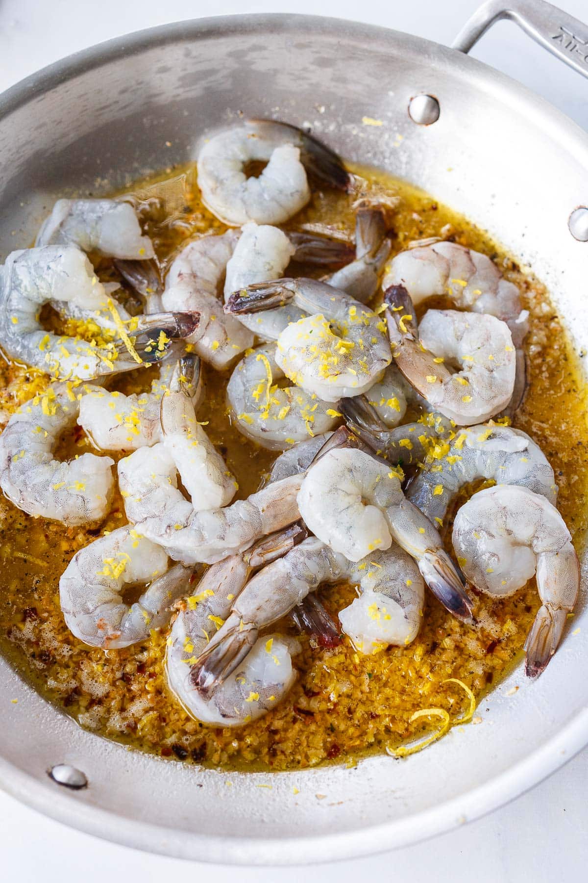 skillet with uncooked shrimp tossed in pan of garlic butter sauce and lemon zest.