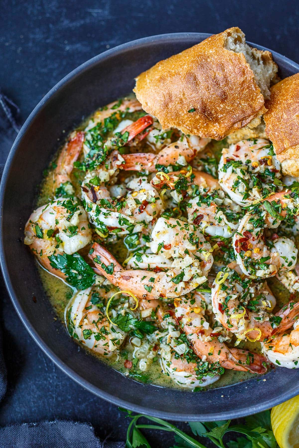 plate of shrimp scampi garnished with parsley and lemon zest, with two pieces of crusty bread to dip in rich sauce.