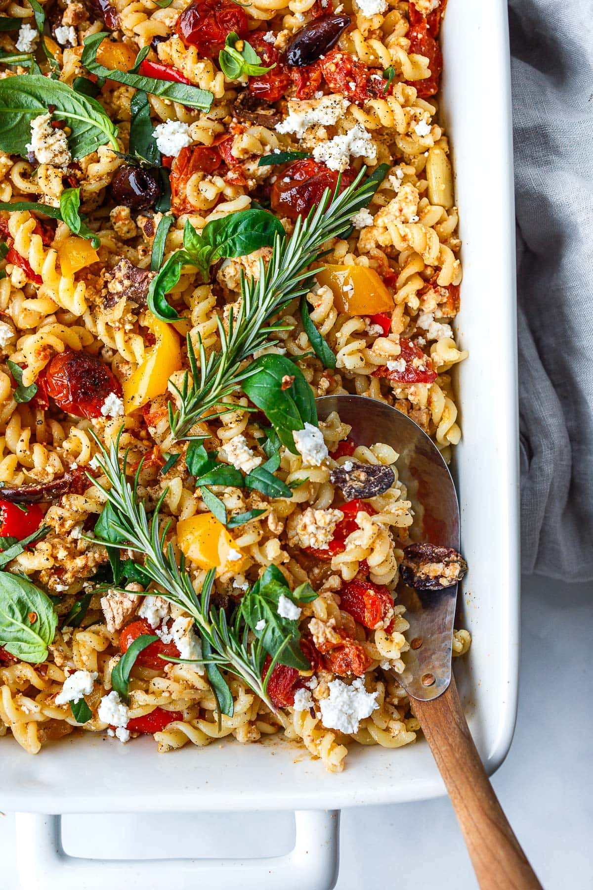 rectangular dish with baked feta pasta with tomatoes and olives and fresh herbs and a wood and metal serving utensil. 