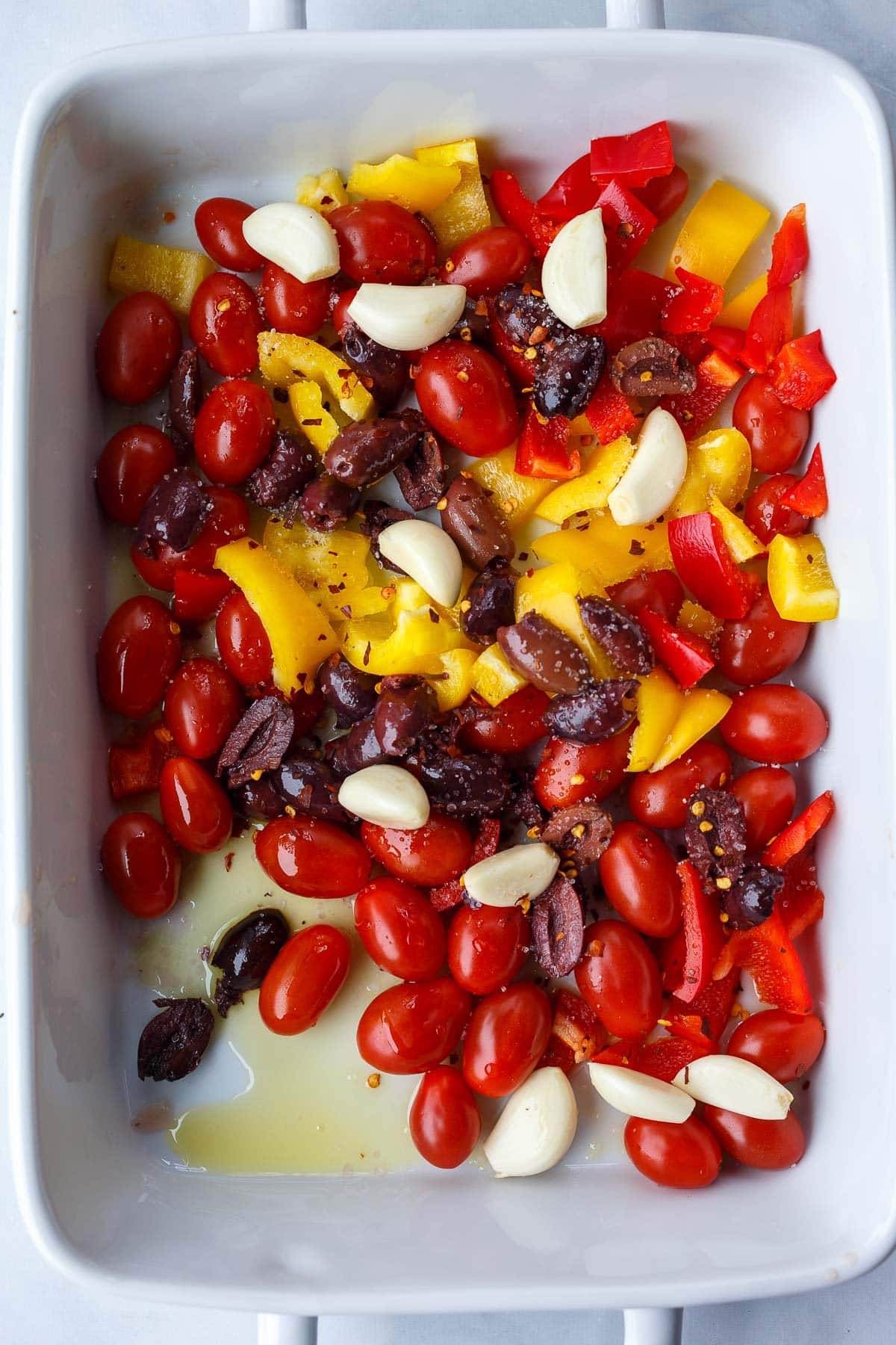 rectangular baking dish with fresh tomatoes, bell peppers, olives, garlic cloves, red pepper flakes, salt, pepper, and olive oil. 