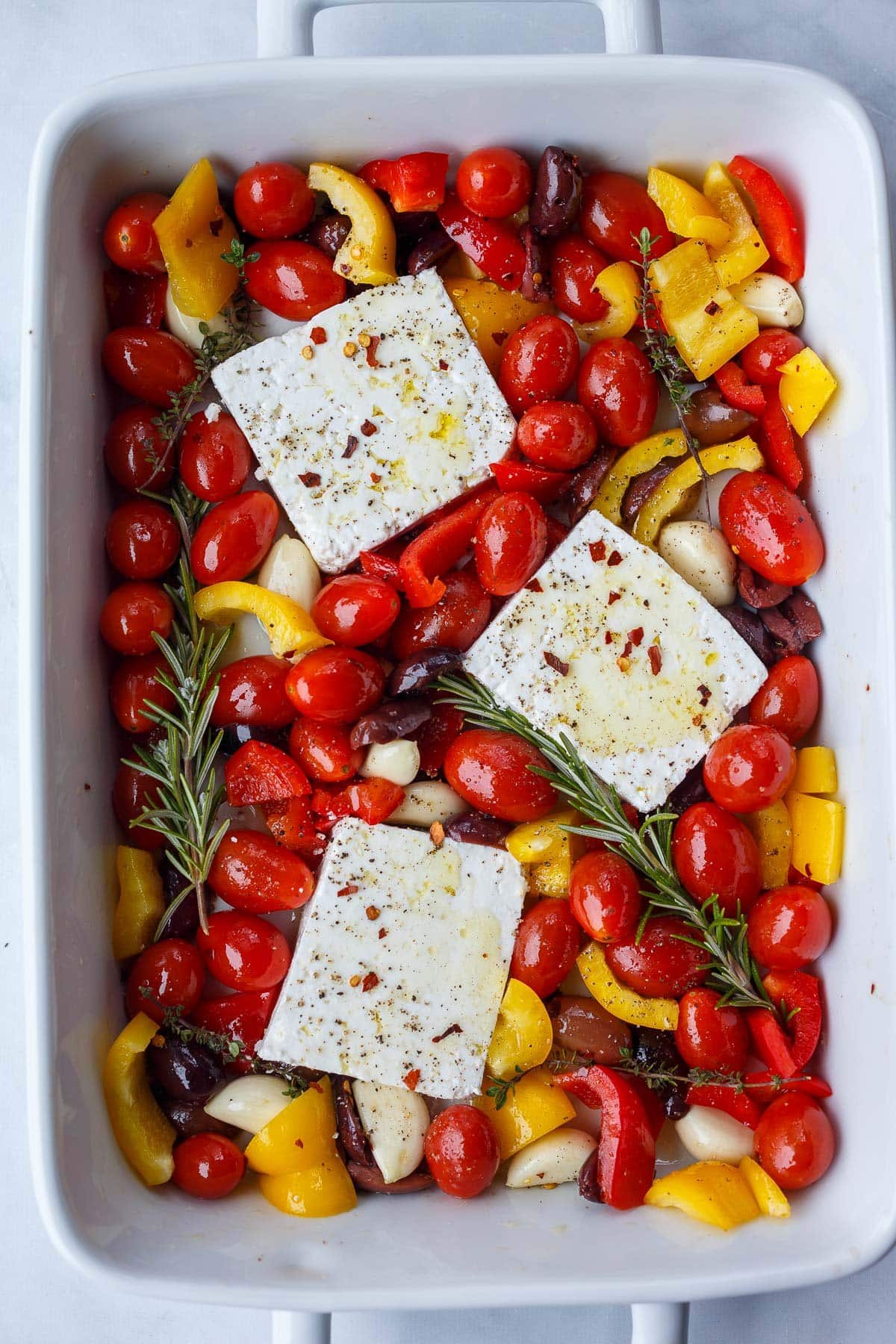 rectangular baking dish with three blocks of feta nestled in between tomatoes, peppers, olives, and rosemary sprigs. 