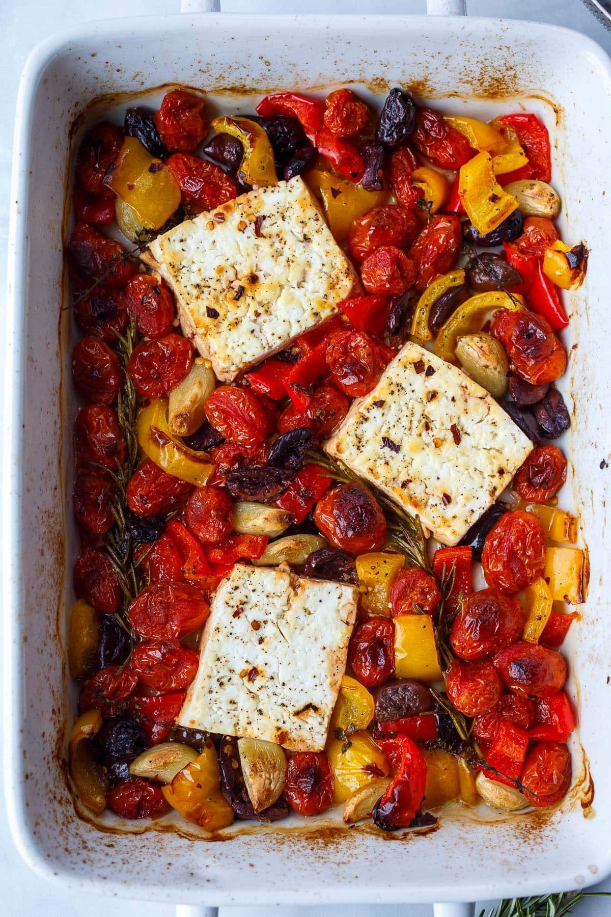 baked feta blocks in a rectangular baking dish with baked and charred vegetables, olives, and herbs. 