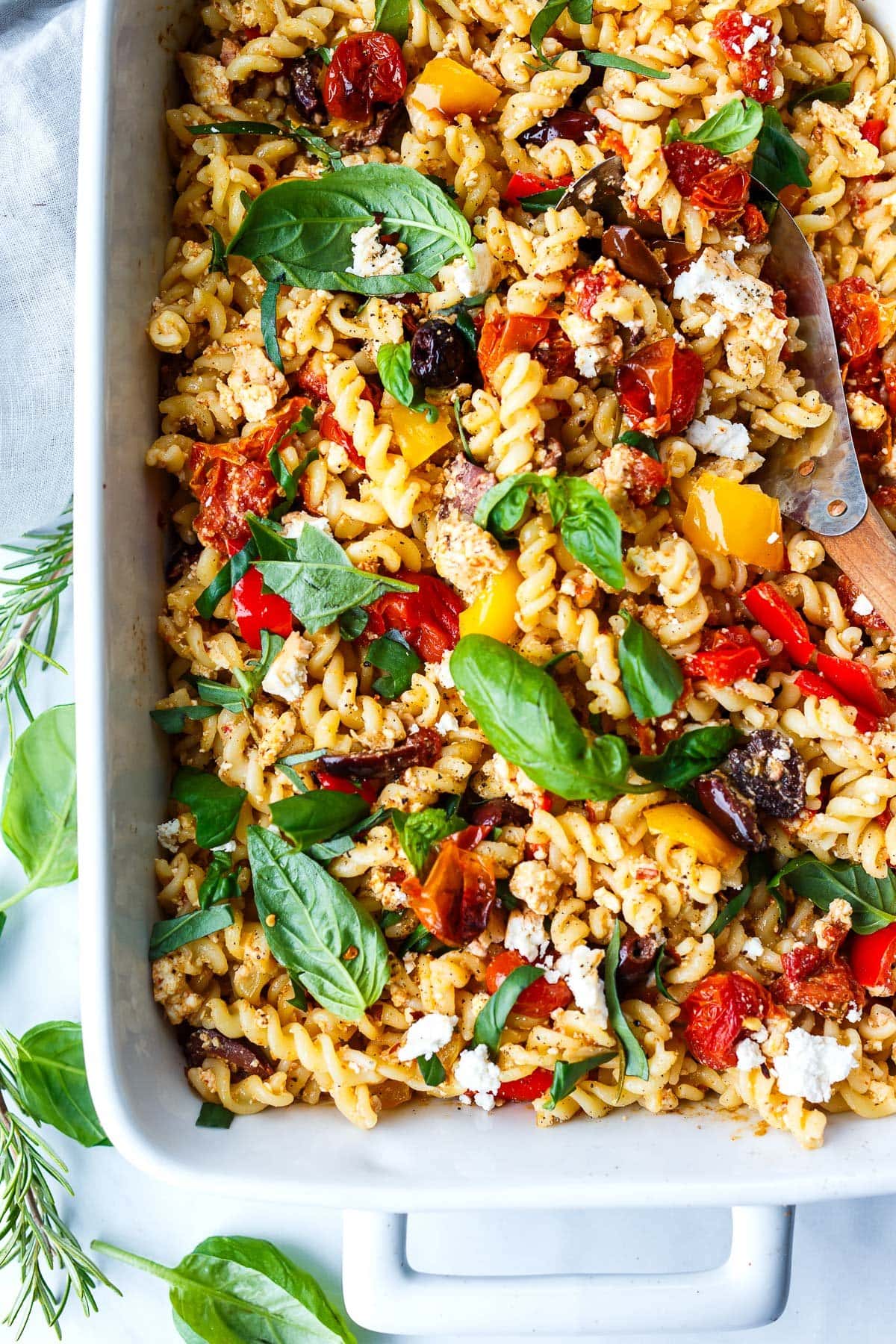 rectangular dish with baked feta pasta with tomatoes and olives and fresh herbs and a wood and metal serving utensil. 