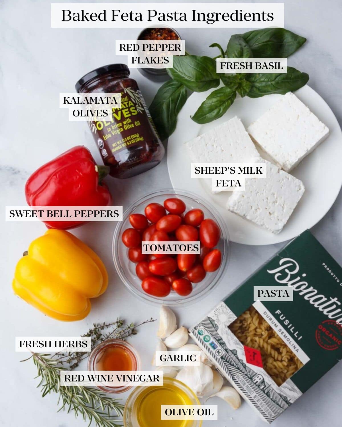 Baked Feta Pasta Ingredients arranged and labeled on a counter - red pepper flakes, fresh basil, kalamata olives, sheep's milk feta, sweet bell peppers, tomatoes, pasta, garlic, fresh herbs, red wine vinegar, olive oil. 