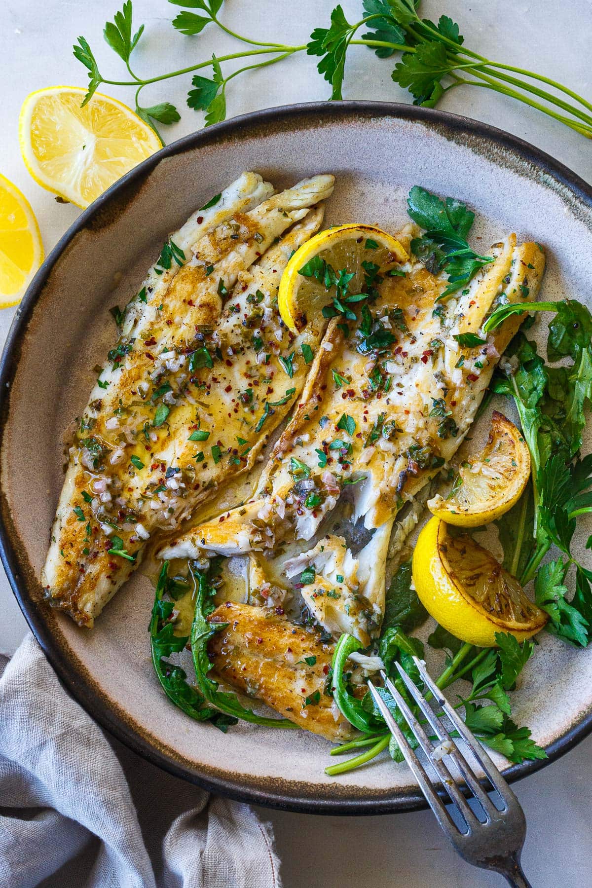 pan seared branzino fillets on a plate with a fork showing the flaky texture, topped with lemon butter sauce, fresh herbs, and seared lemon wedges.