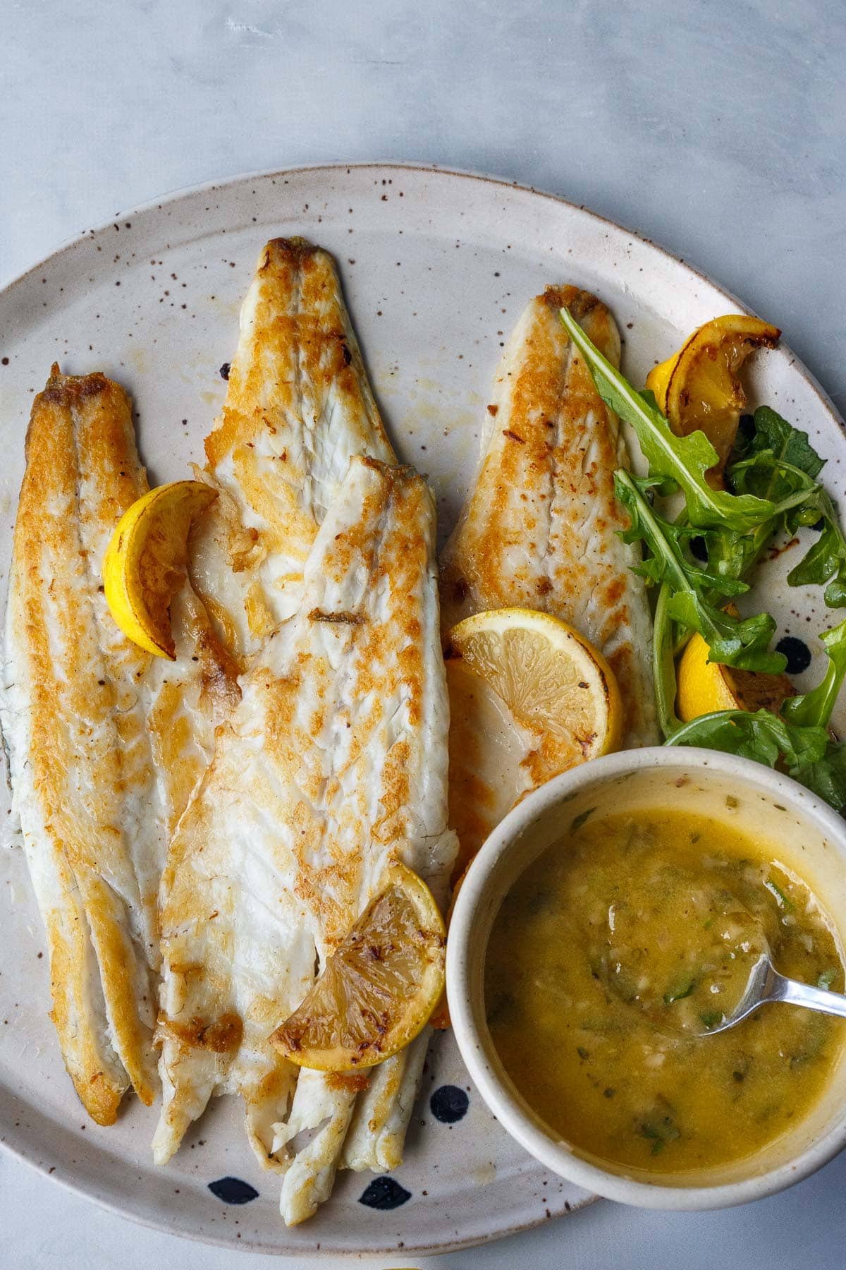 pan seared branzino fillets on a plate with lemon wedges and arugula, and a small bowl of lemon butter sauce with a spoon.