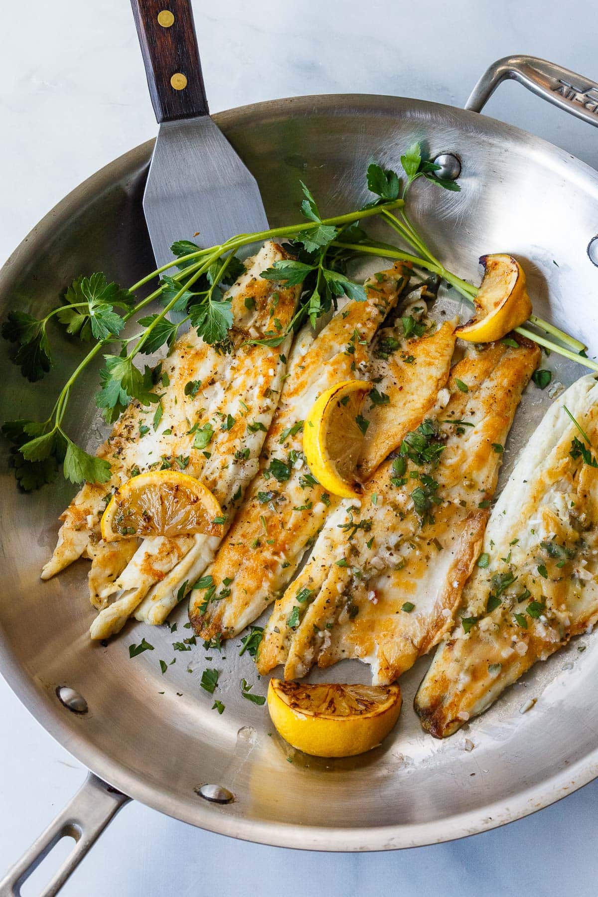 pan seared branzino fillets in a skillet with seared lemon wedges and sprigs of parsley with lemon butter sauce over top of the fillets.
