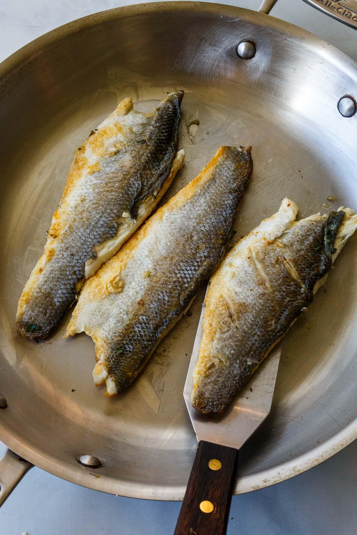 three branzino fillets searing in a skillet with a metal spatula beginning to flip one.