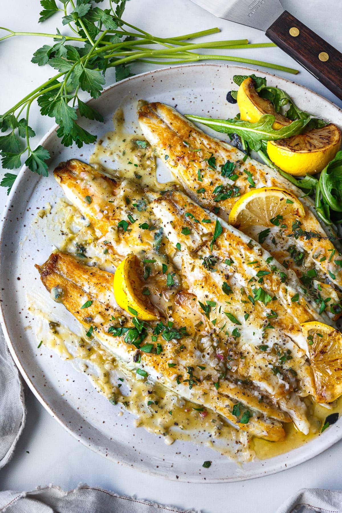 pan seared branzino on plate topped with lemon butter sauce, fresh herbs, and lemon wedges.