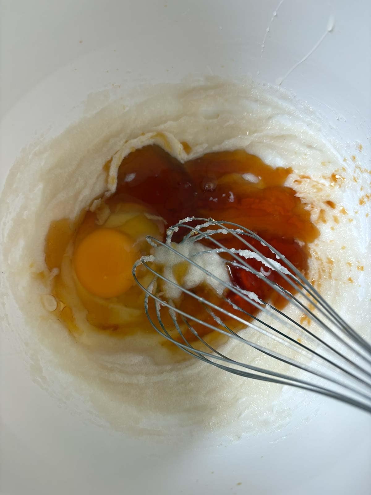 mixing bowl with melted coconut and sugar mixture with maple syrup and egg added and a whisk to mix it up.