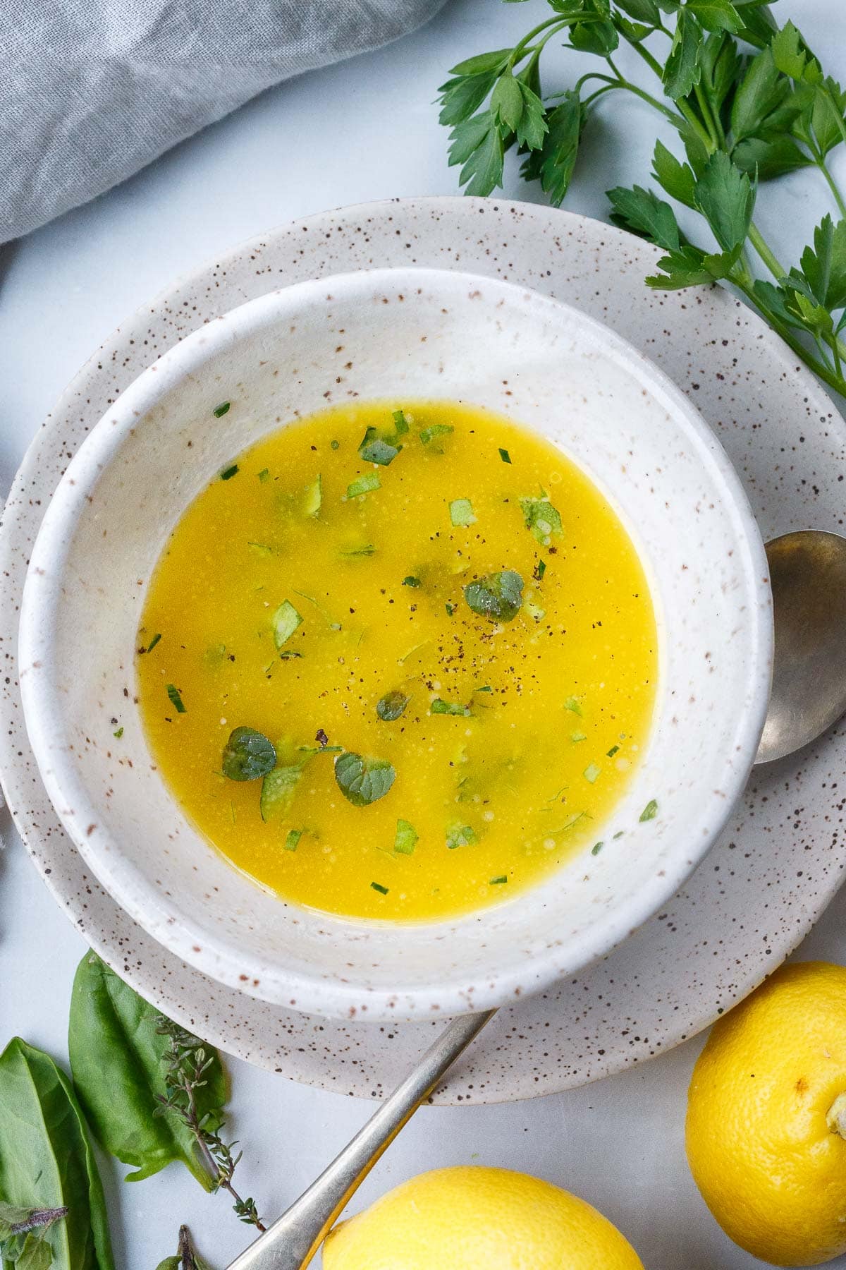small dish of lemon butter sauce on plate with silver spoon next to lemons and fresh herbs. 