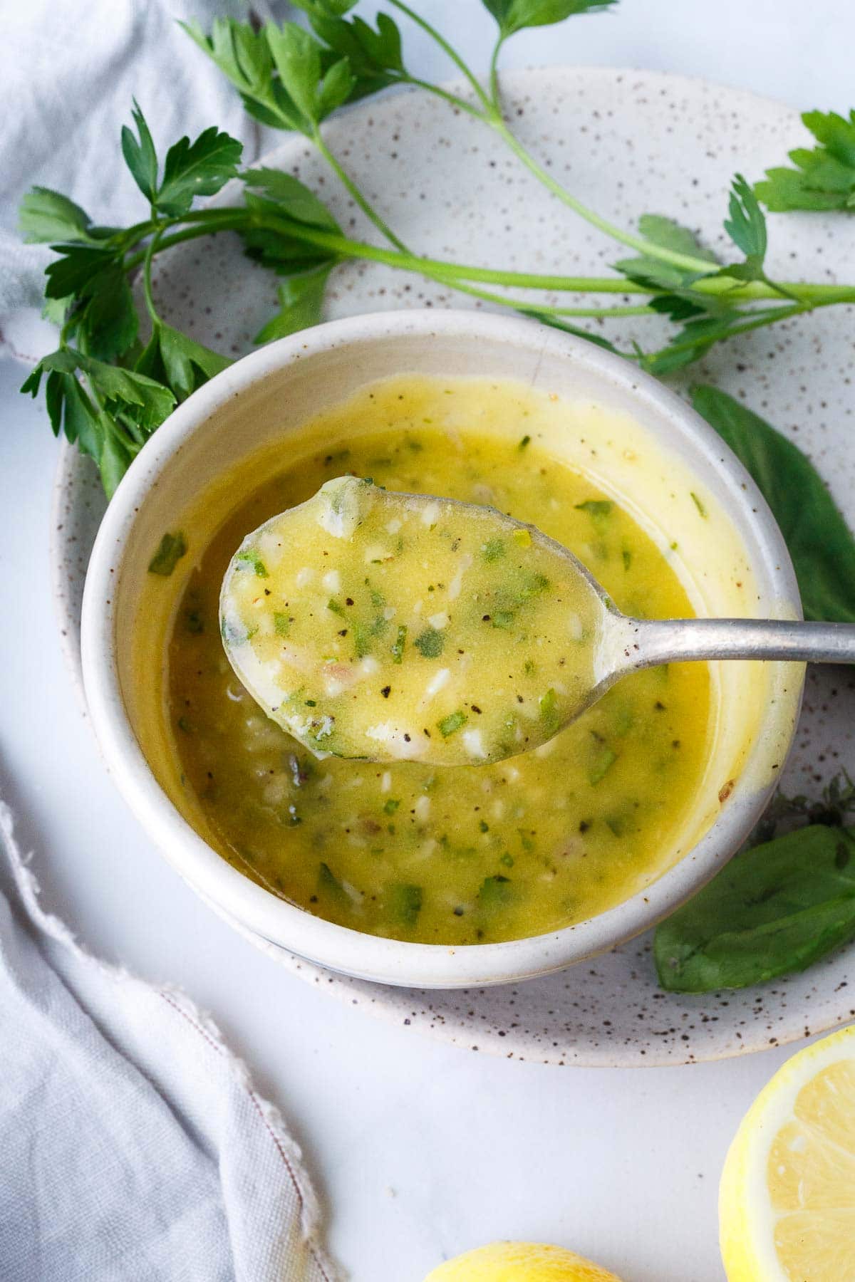 spoon lifting up a spoonful of lemon butter sauce with garlic and herbs and pepper above small bowl.