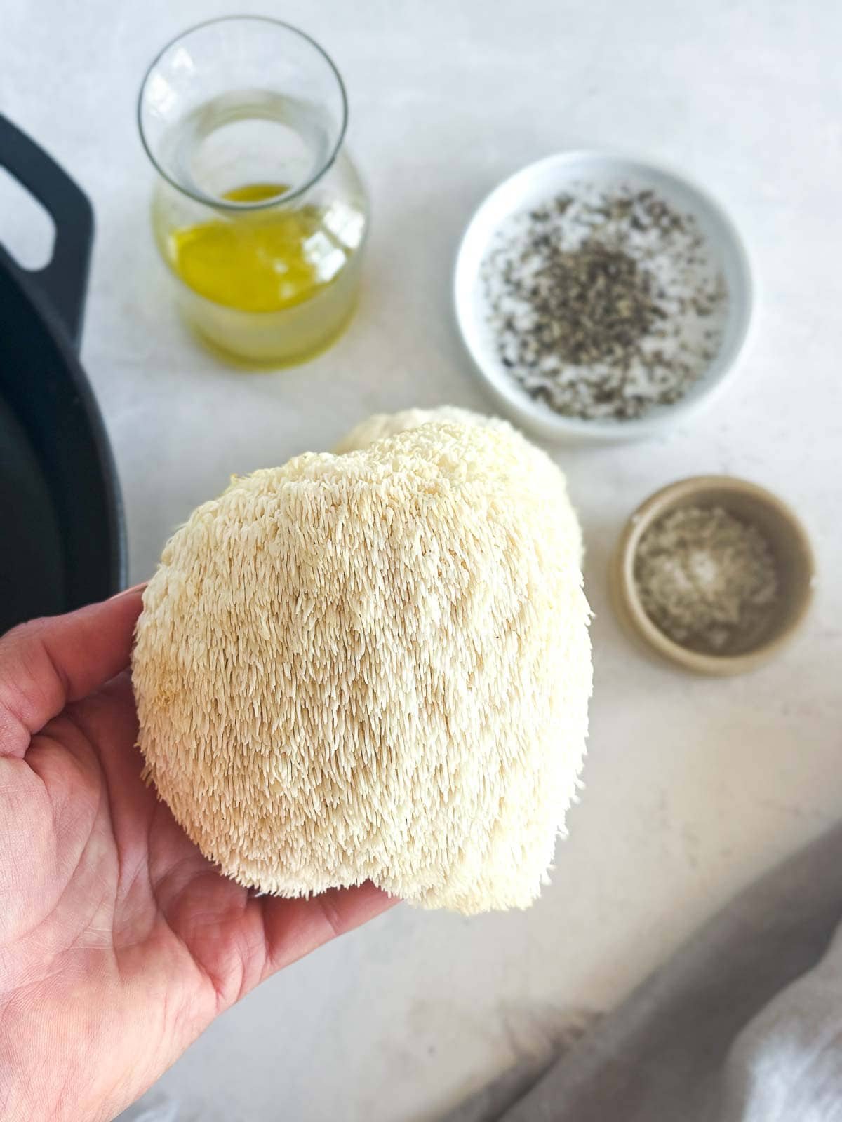 hand holding up fresh lion's mane mushroom with jar of oil and small bowls of spices in background.