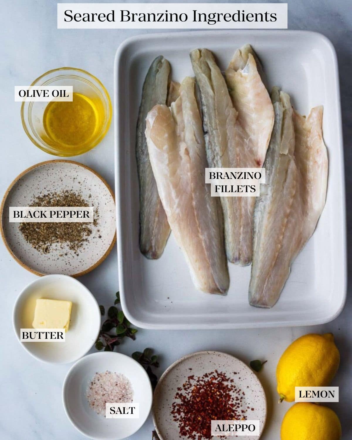 branzino recipe ingredients labeled with branzino fillets in rectangular dish, olive oil in a bowl, black pepper, butter, salt, and Aleppo in small dishes and two lemons.