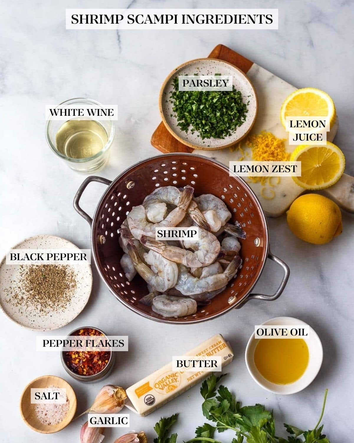 shrimp scampi ingredients- shrimp in colander, next to glass of white wine, minced parsley, lemon zest and cut lemons, spices, small bowl of oil, stick of butter, garlic cloves, and pile of parsley stems.