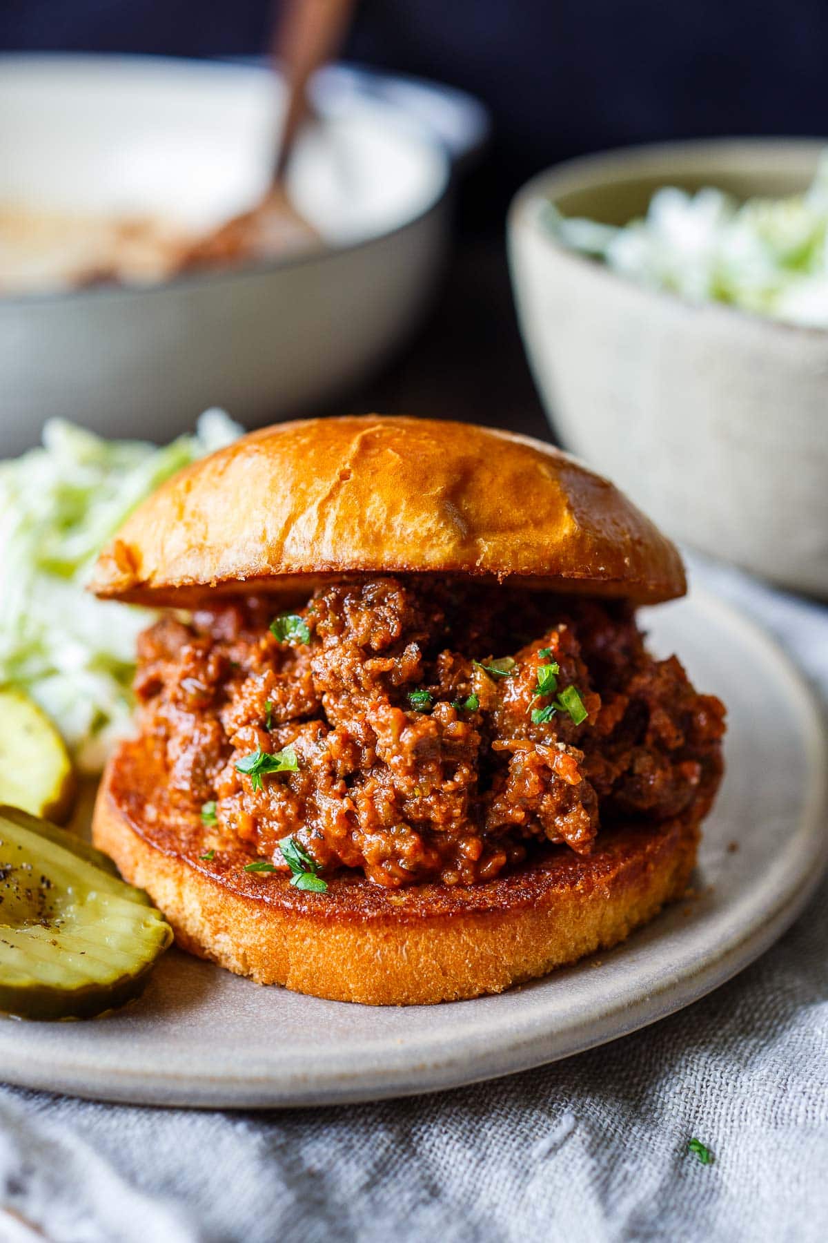 sloppy joes on brioche bun on plate next to pickles. 