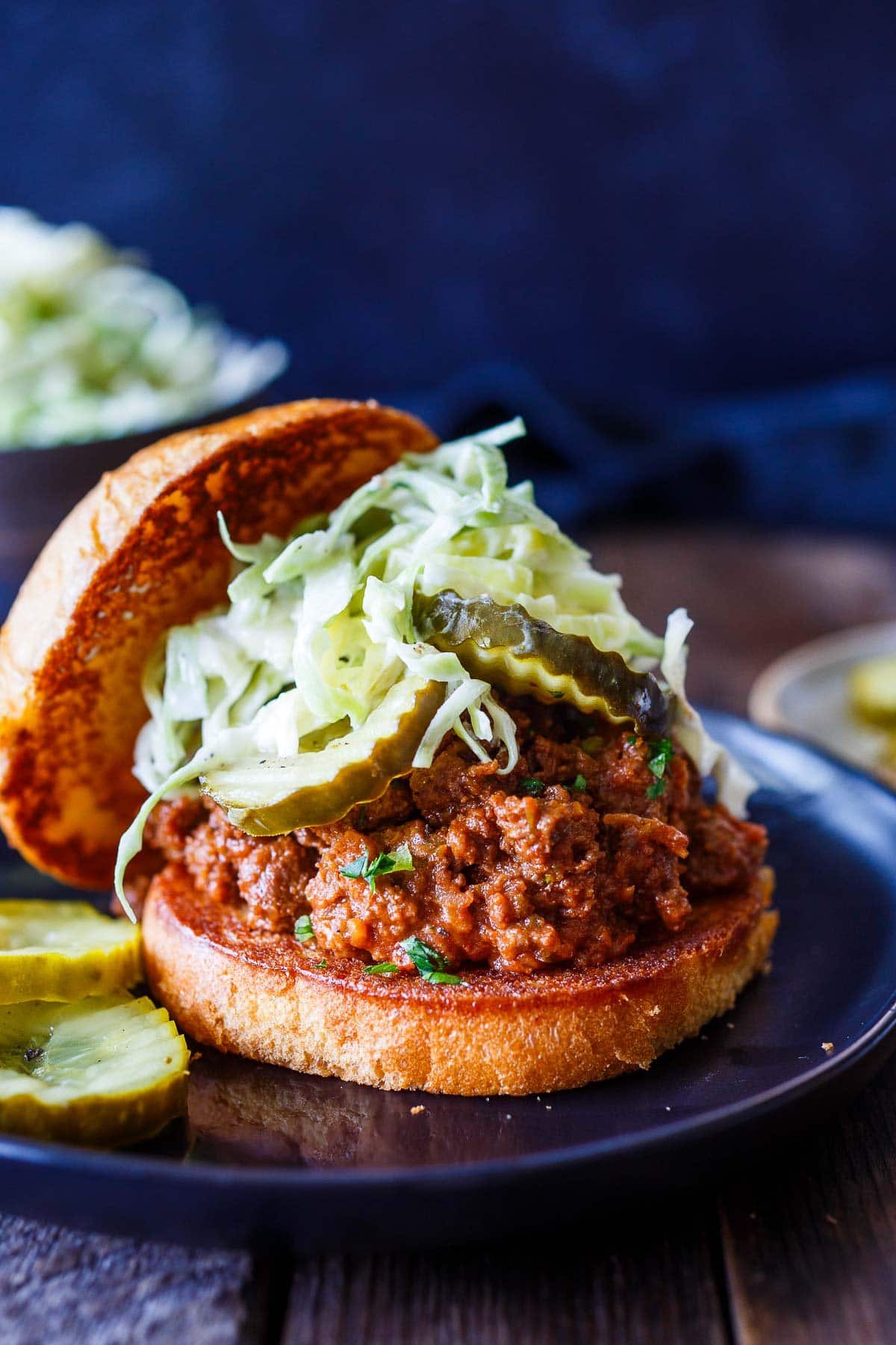 sloppy joes on brioche bun topped with pickles and coleslaw with top bun leaning against the side on plate with pickles. 