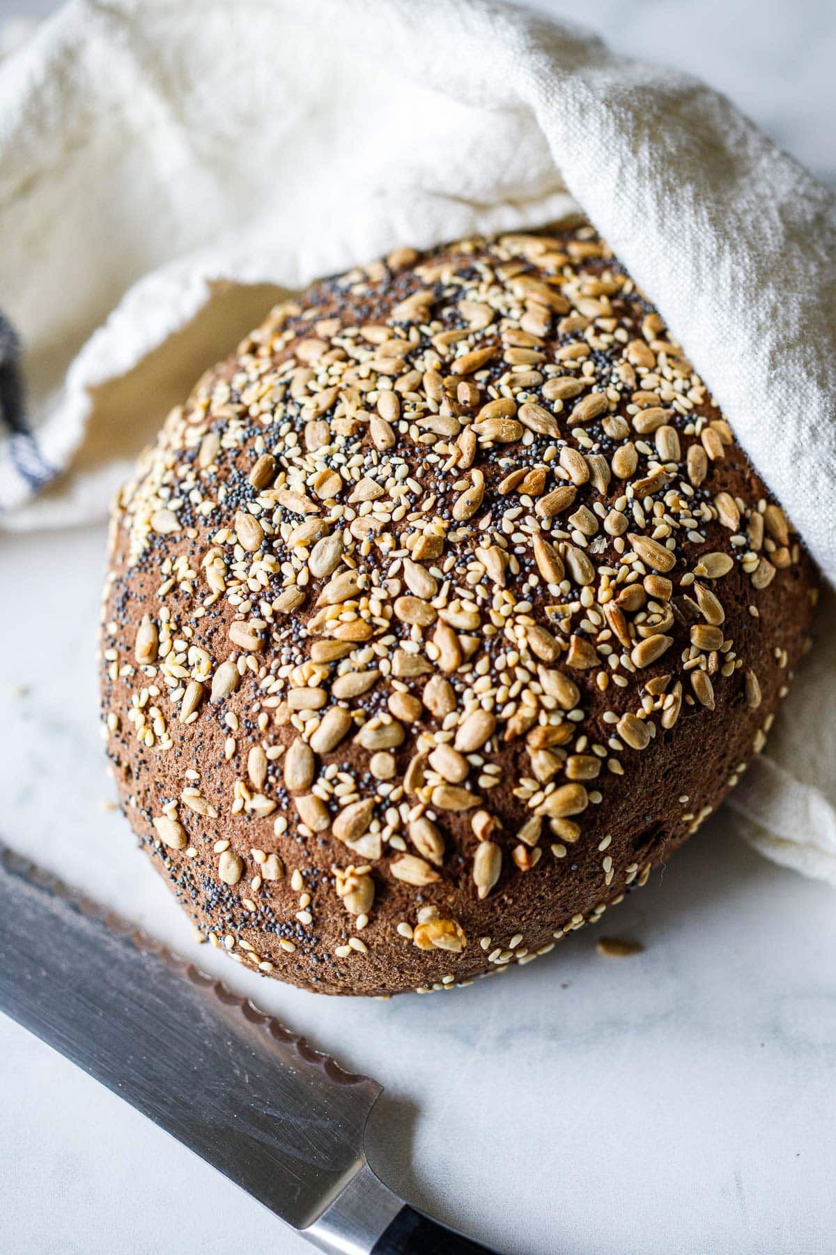 loaf of gluten free bread with seeds on top partially wrapped in linen towel next to serrated knife.