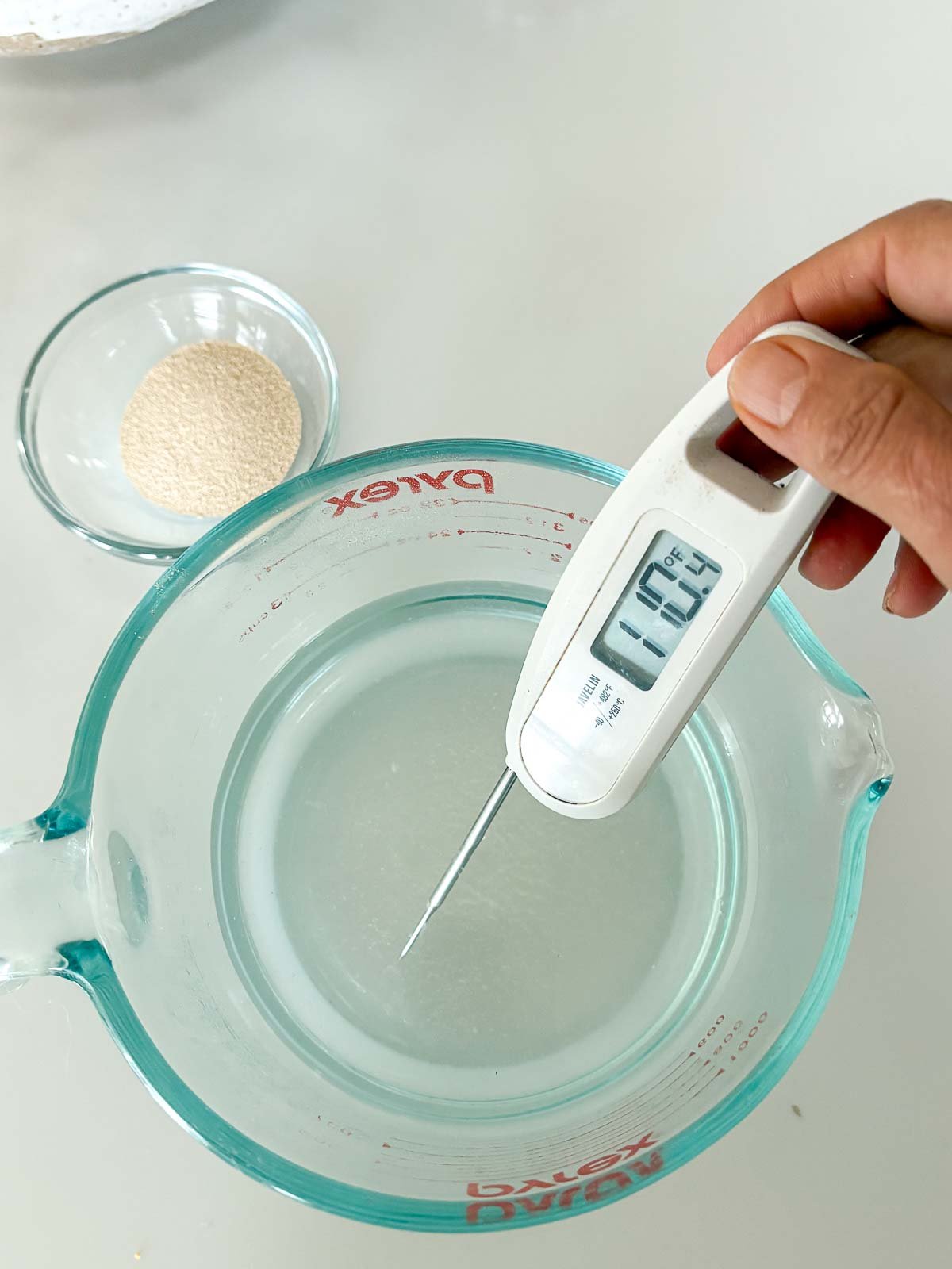 holding thermometer in measuring cup of water reading 110 degrees next to small bowl of yeast.