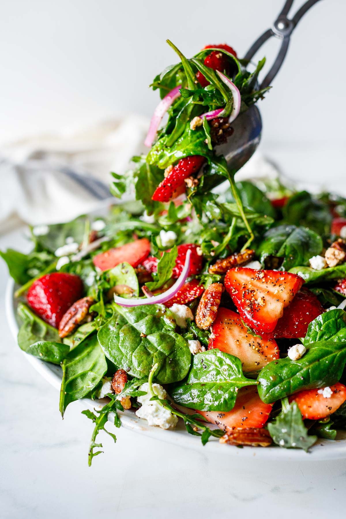 strawberry and spinach salad with salad tongs picking up the salad, full of pecans, goat cheese, onions, and poppy seed dressing.