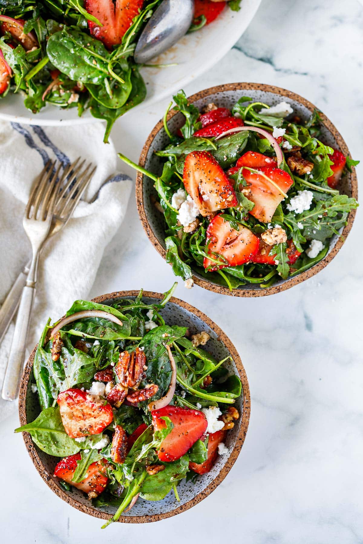two salad bowls with strawberry and spinach salad with pecan brittle, goat cheese, red onion, and balsamic poppy seed dressing, next to forks and a larger salad bowl.
