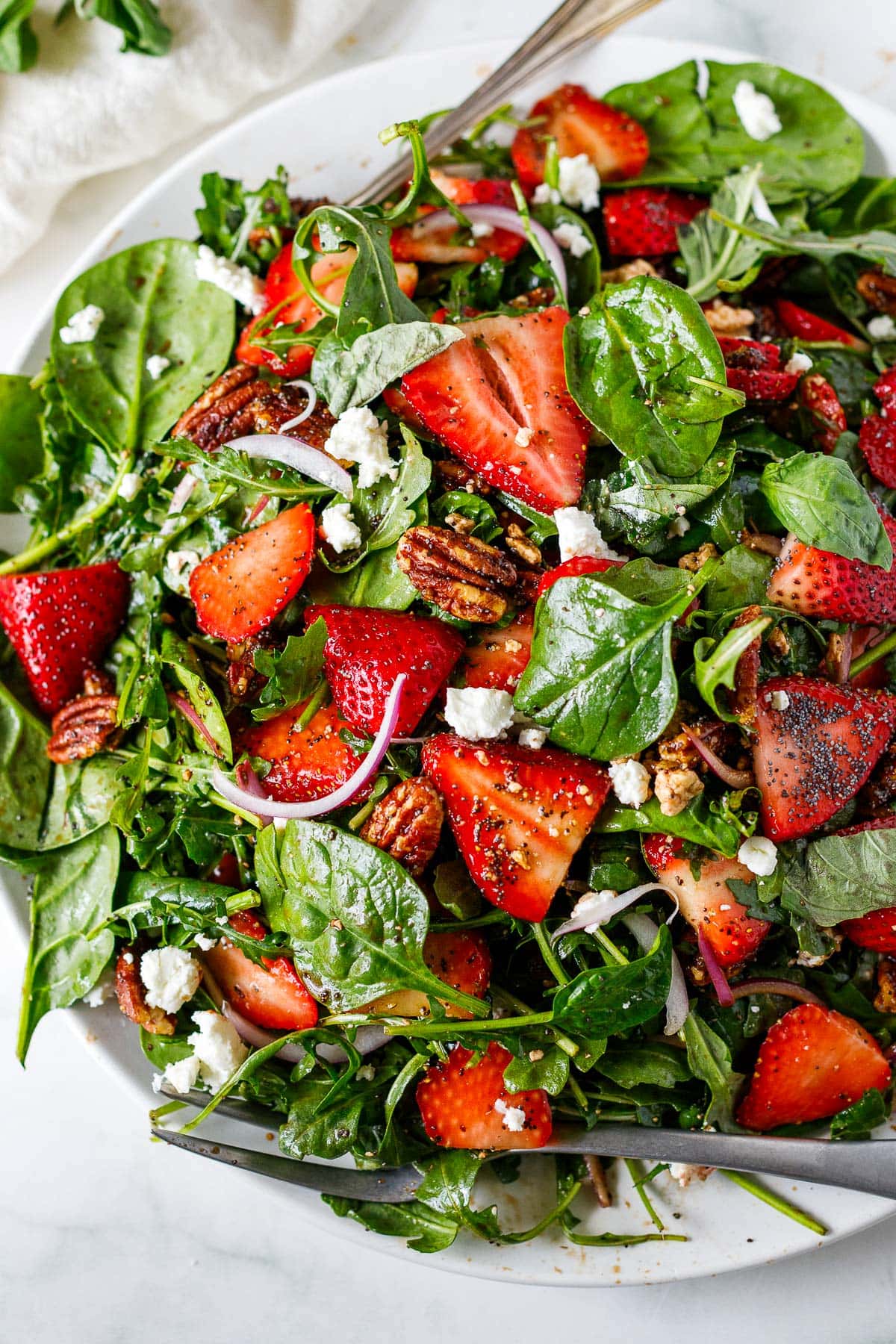 strawberry and spinach salad in large bowl with serving utensils served with goat cheese, pecan brittle, and red onion.