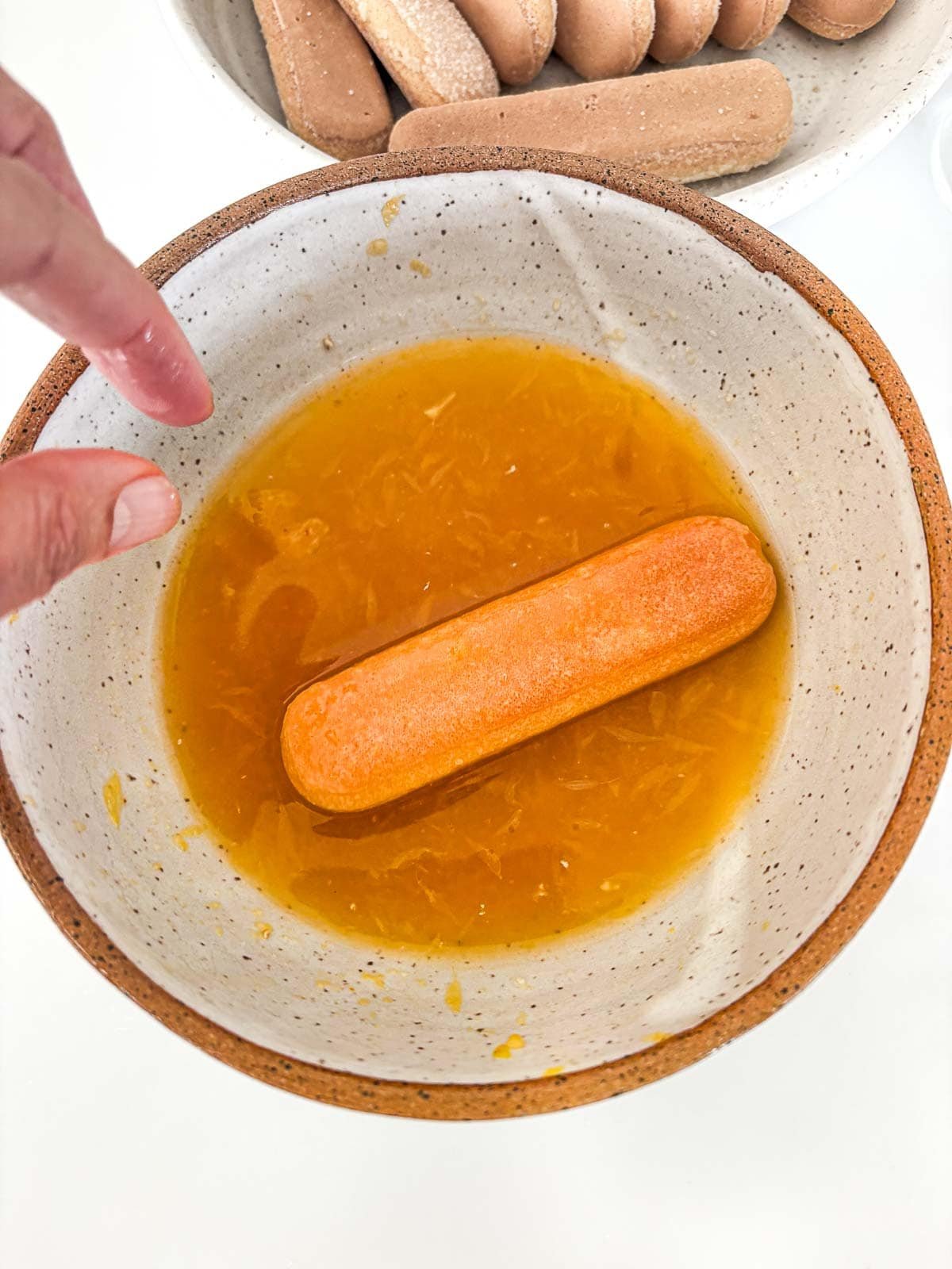 fingers dipping a ladyfinger in the bowl with the orange juice mixture.