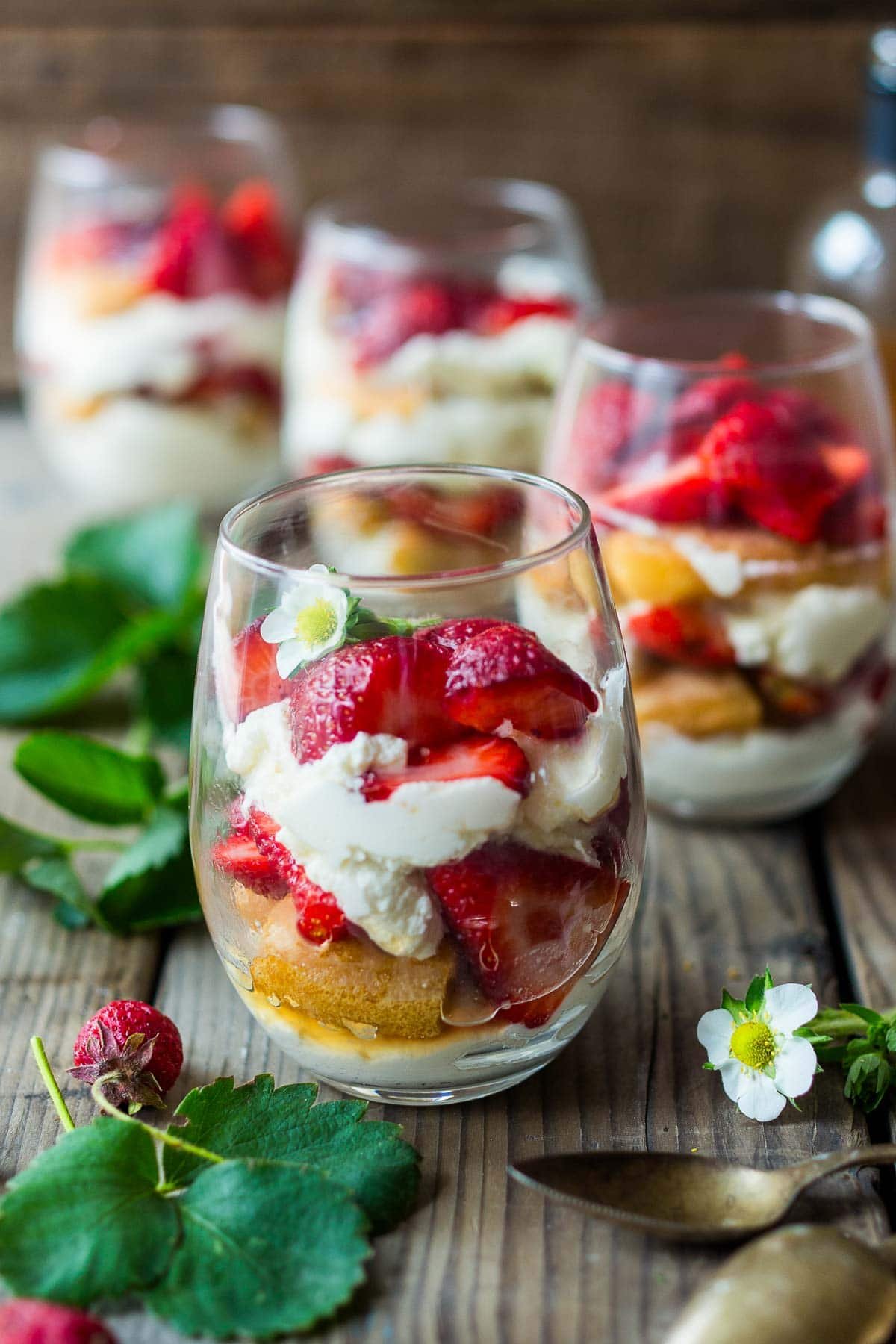 strawberry tiramisu in glasses with layers of mascarpone, ladyfingers, and strawberries, topped with white edible flower on wood surface with spoons and herbs.