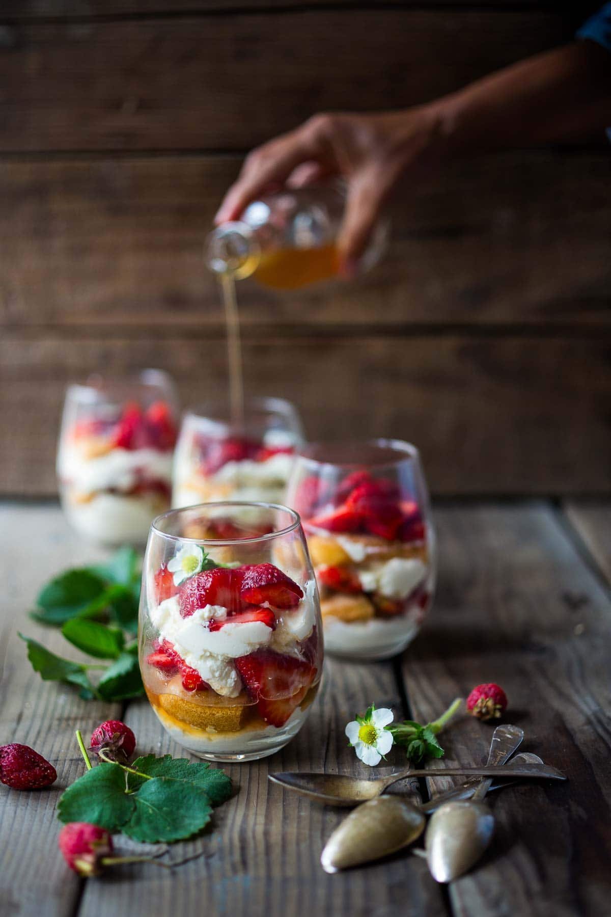 four glasses of strawberry tiramisu on a wood surface with hand pouring elderflower syrup over top of one of the glasses in the back.