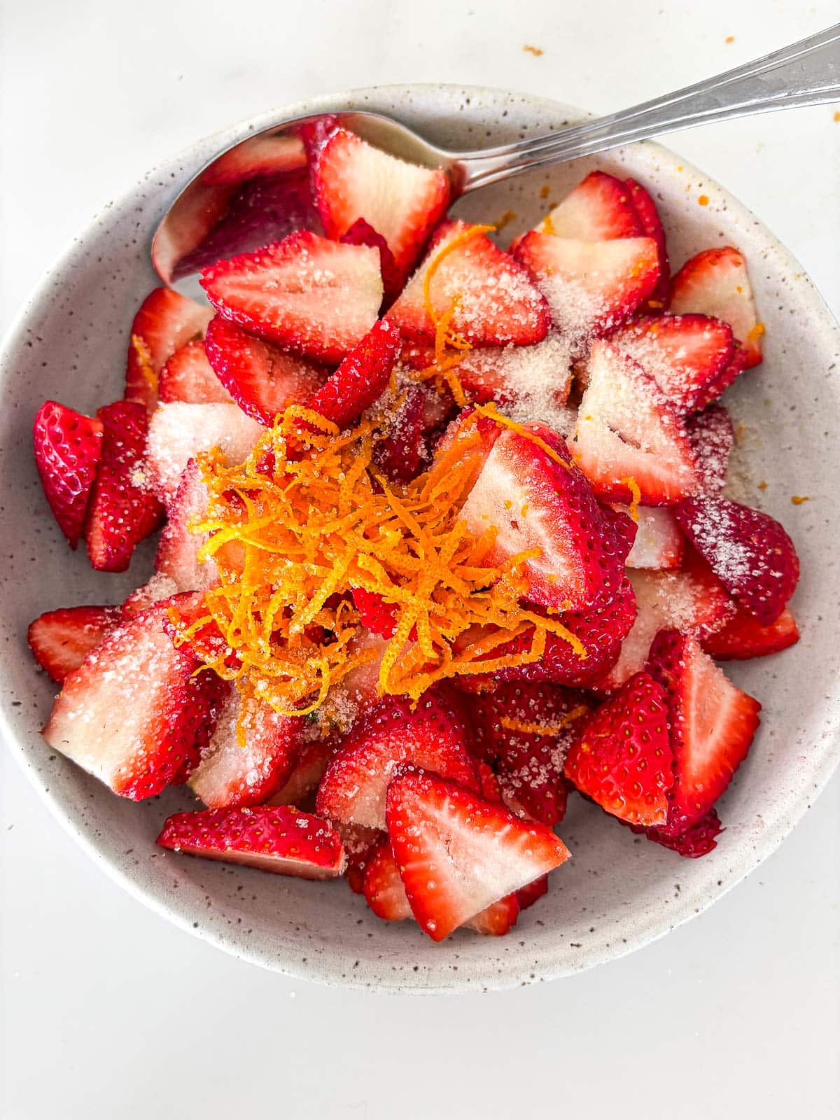 bowl of halved strawberries with sugar and orange zest in bowl with spoon.