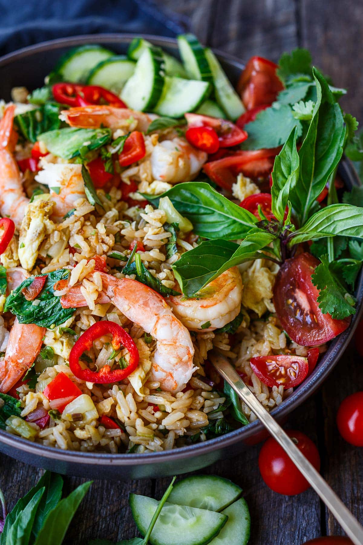 bowl of thai fried rice with shrimp served with a spoon, garnished with fresh herbs, sliced chilies, tomatoes, and cucumbers. 