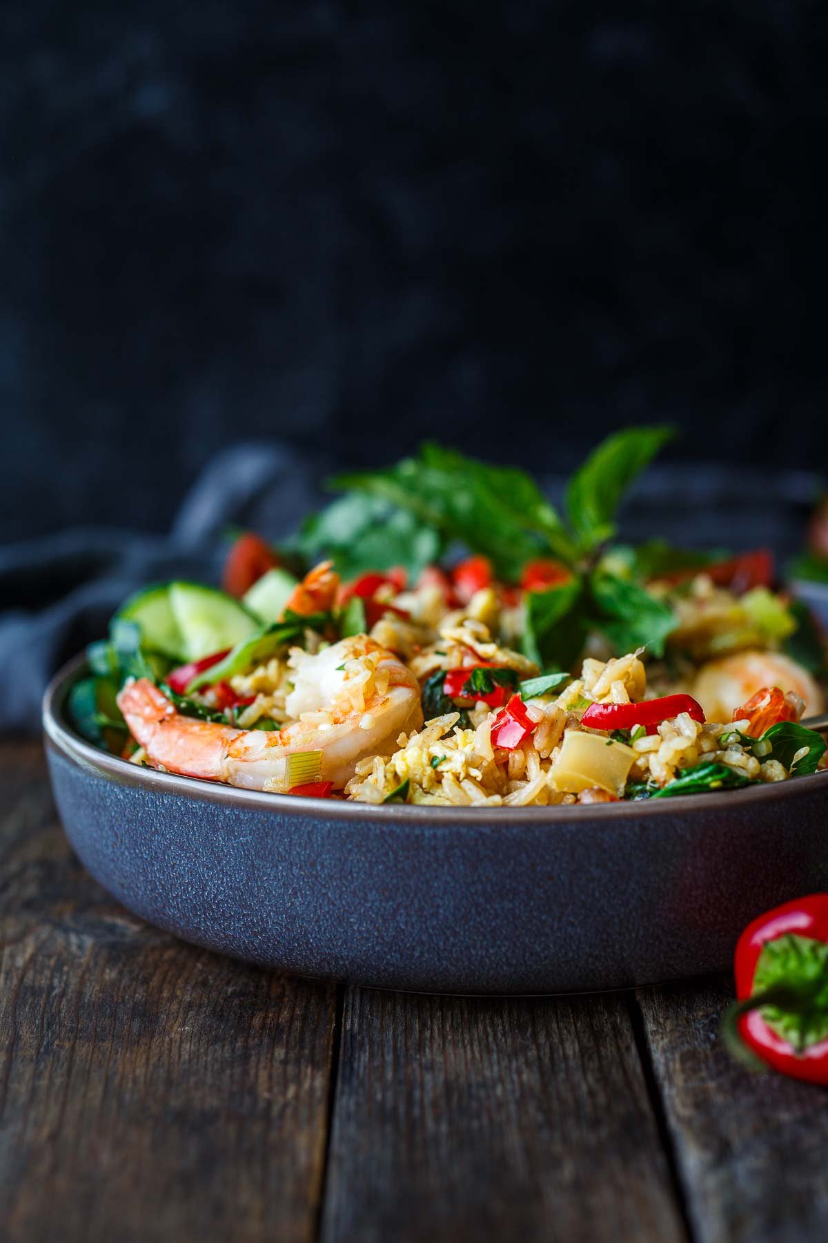 bowl of thai fried rice with shrimp with chilies and fresh herbs on wood surface next to a whole chili.