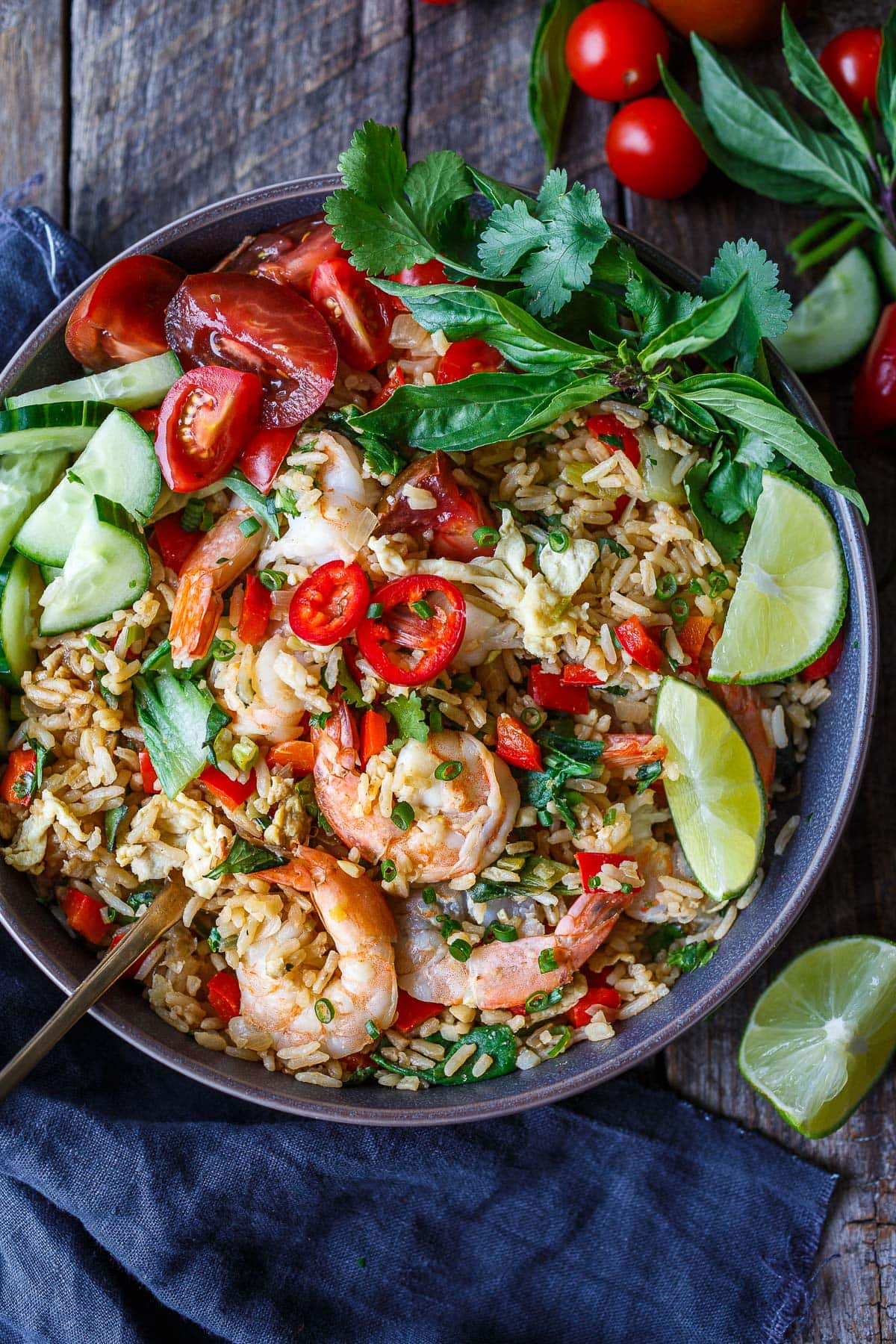 bowl of Thai fried rice with shrimp garnished with lime wedges, chilies, tomatoes, cucumbers, and thai basil.