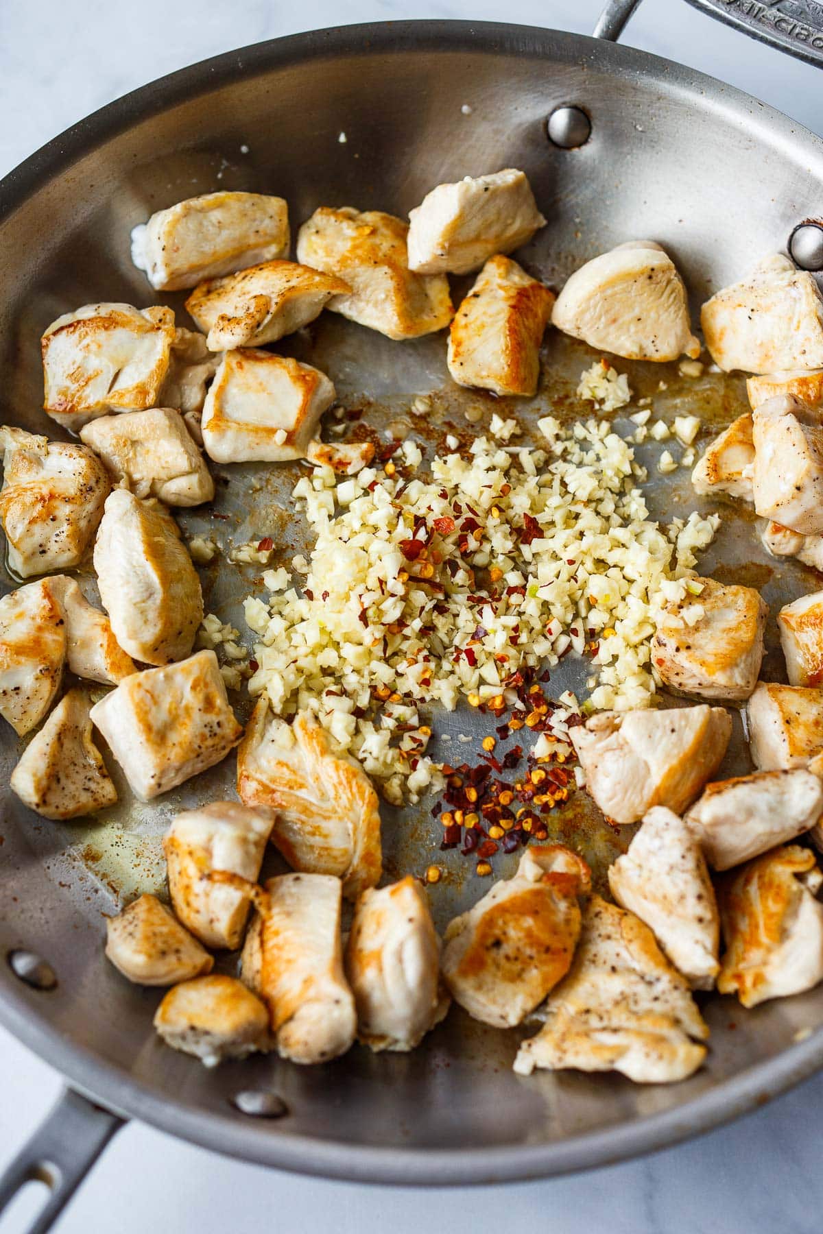 stainless steel skillet with seared bite-size chicken breast surrounded center pile of minced garlic and red pepper flakes.