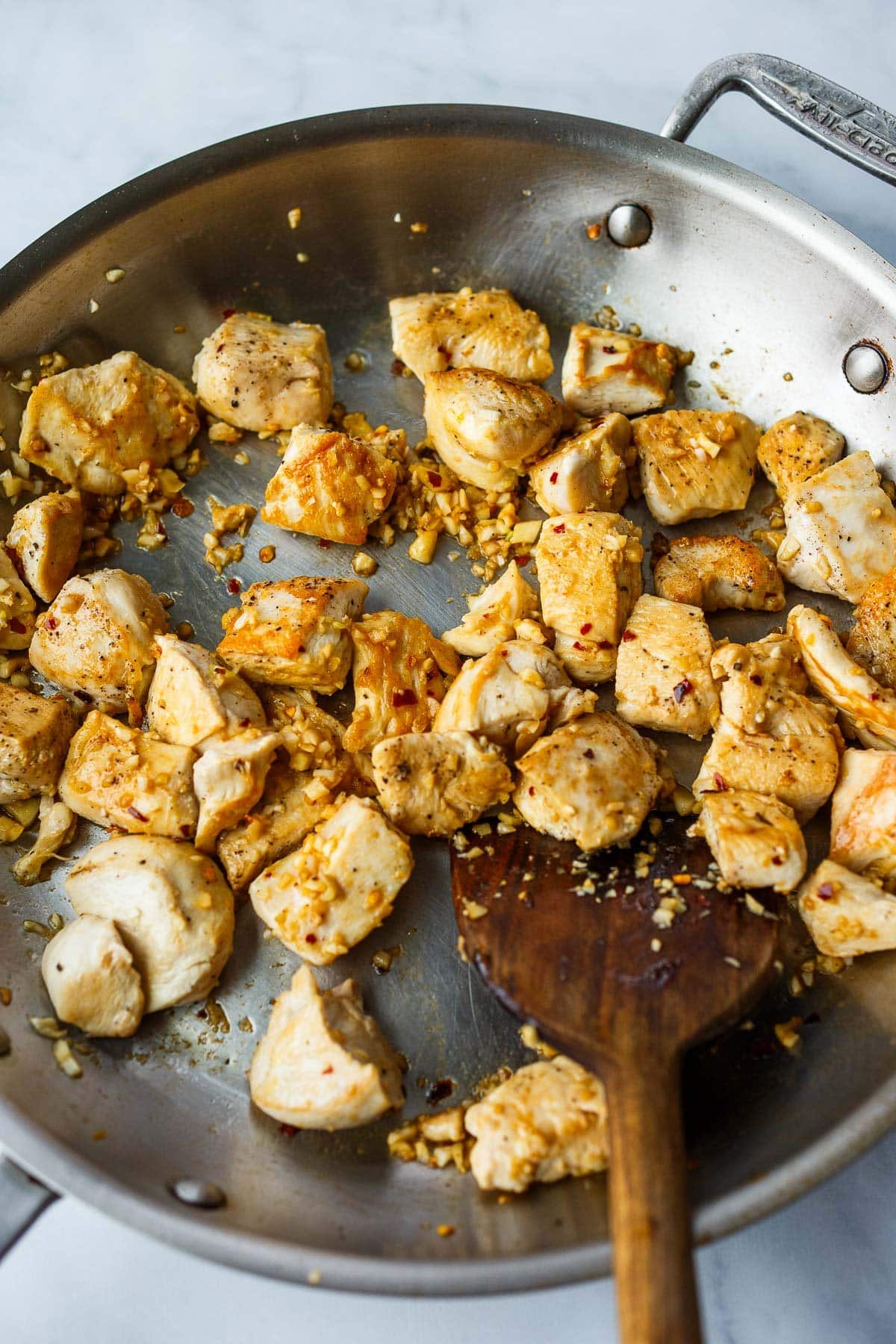 large skillet with seared chicken breast pieces and garlic with wood spoon for stirring.