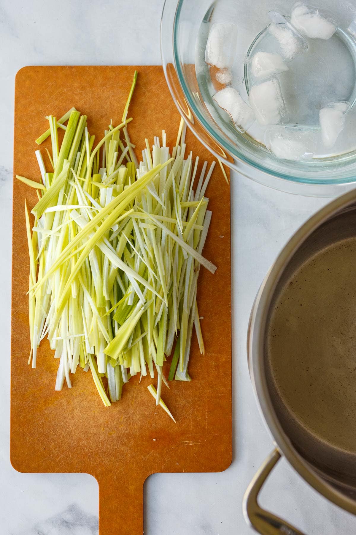 cutting board with light green part of leeks thinly sliced into strips next to a bowl of ice water.