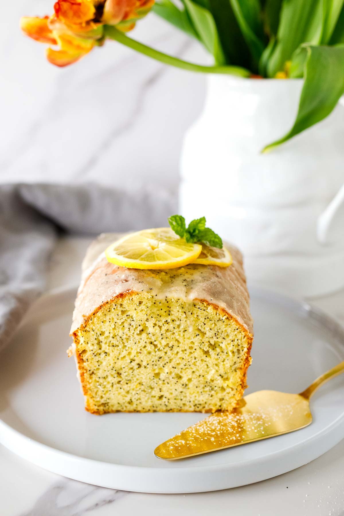 lemon poppy seed cake on plate with gold cake spatula; cake garnished with lemon glaze, lemon slices, and mint leaves. 