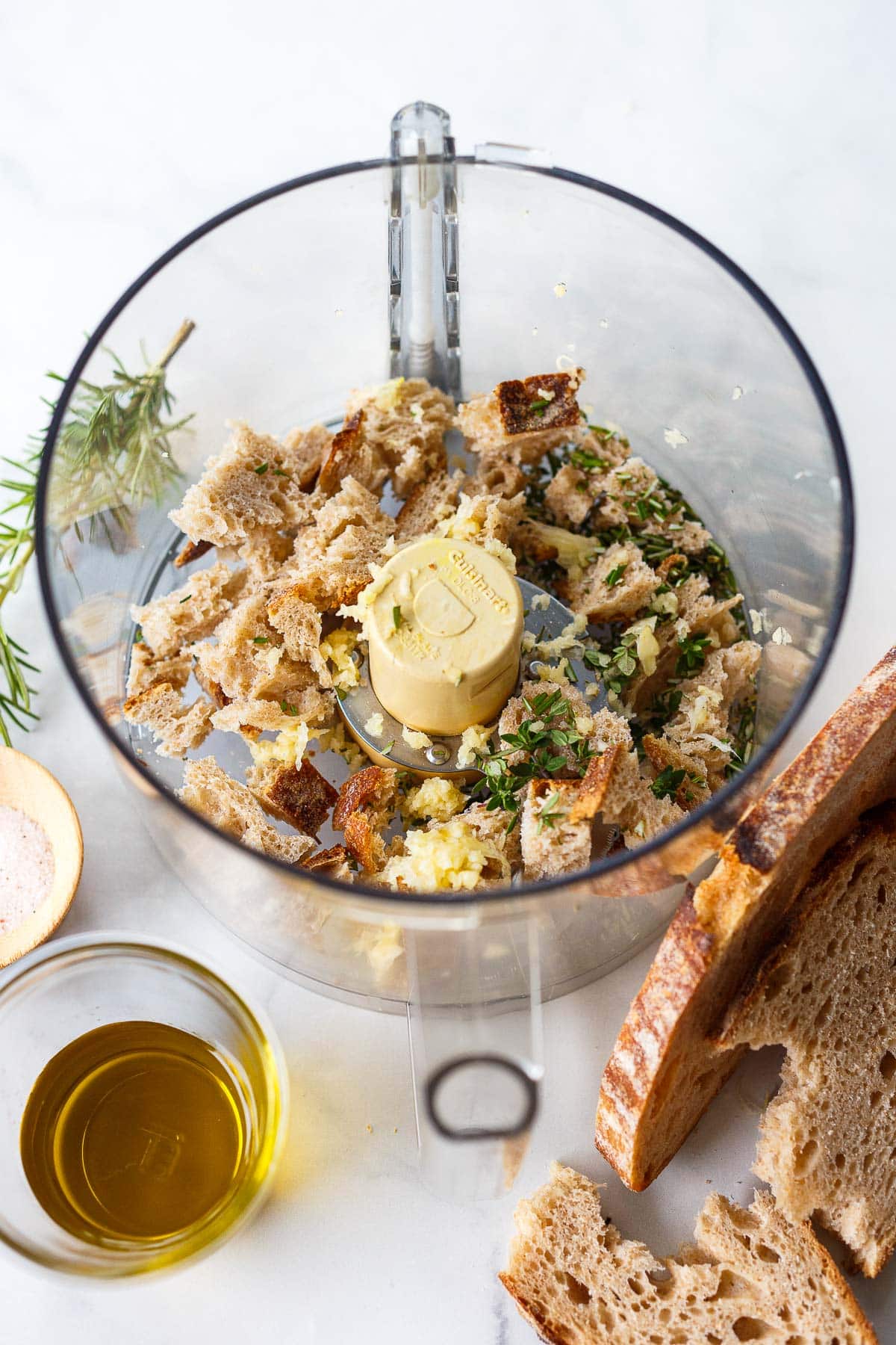 food processor with torn bread, herbs, garlic next to glass of olive oil and more sourdough.