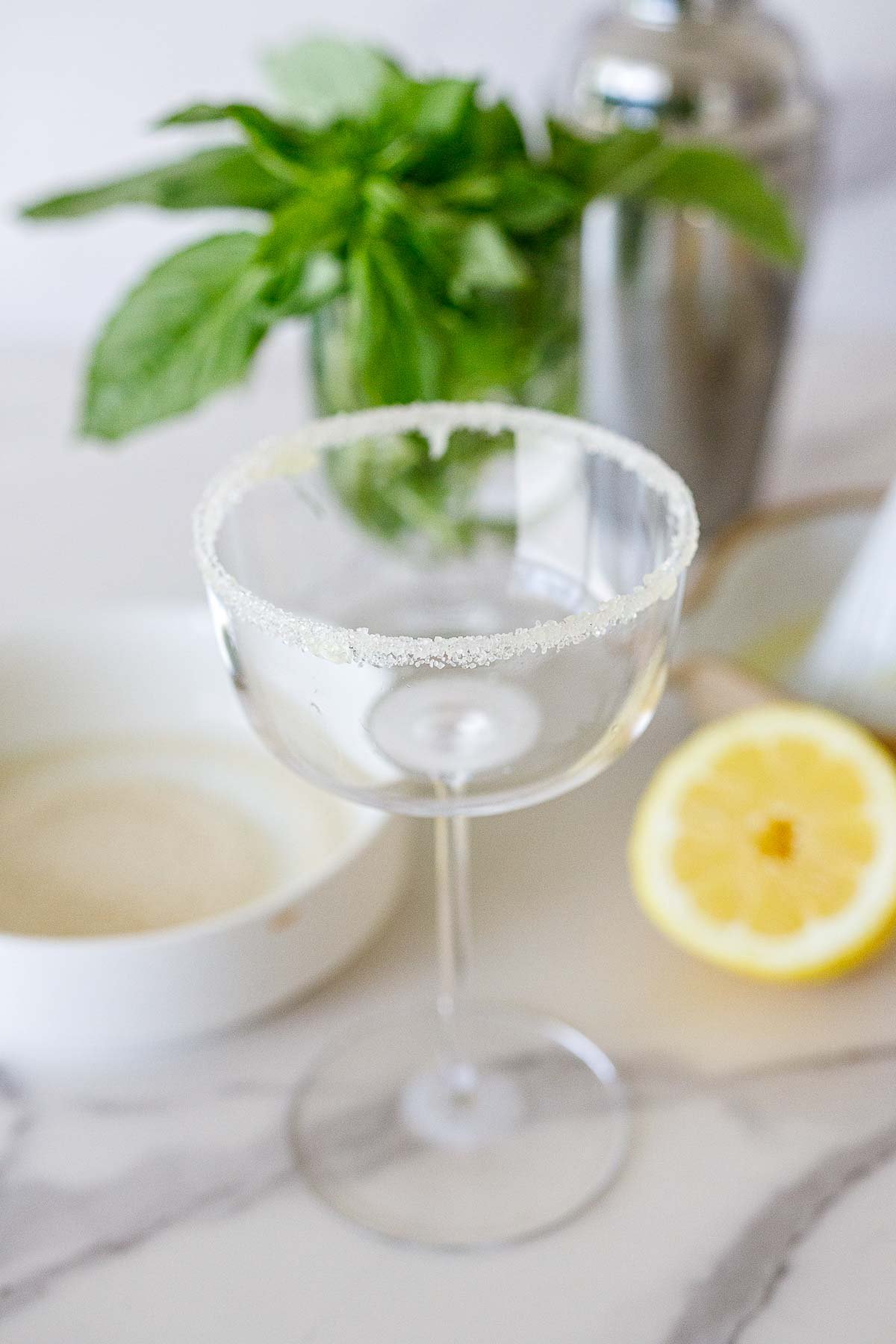 sugar rim on a coupe glass with basil, shaker, and lemon in the background. 