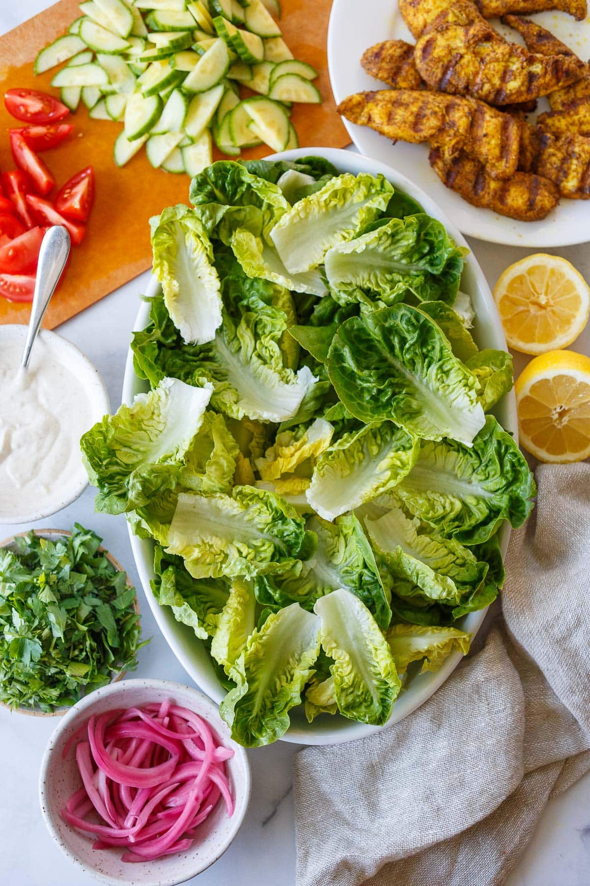 oval salad bowl with little gem lettuce surrounded by prepped ingredients for chicken shawarma salad - pickled onions, cilantro, tahini yogurt dressing, tomato, cucumber, grilled chicken shawarma, and lemon halves.