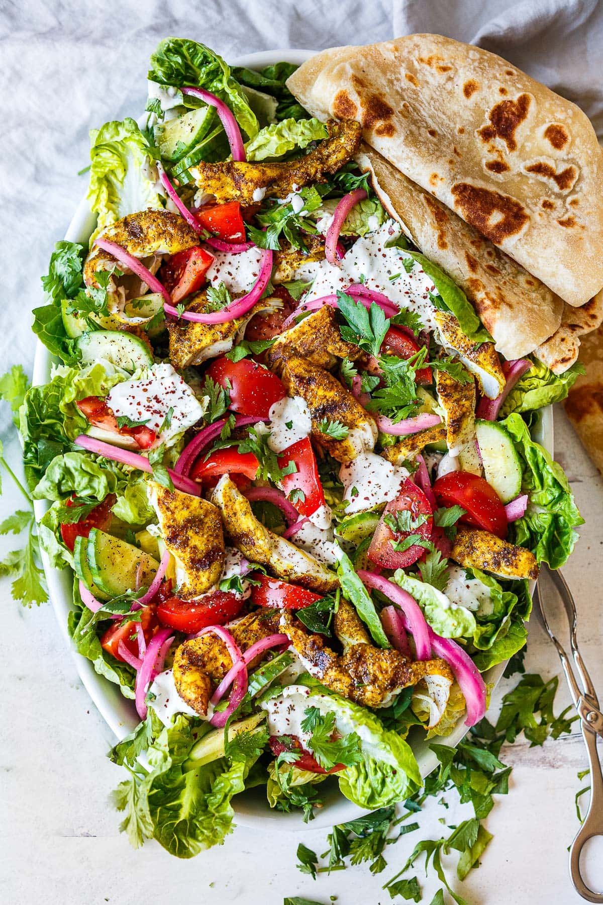 Grilled chicken salad infused with shawarma spice, with little gem lettuce, pickled onions, tomatoes, and cilantro with dollops of tahini yogurt dressing, with pita folded on the side.