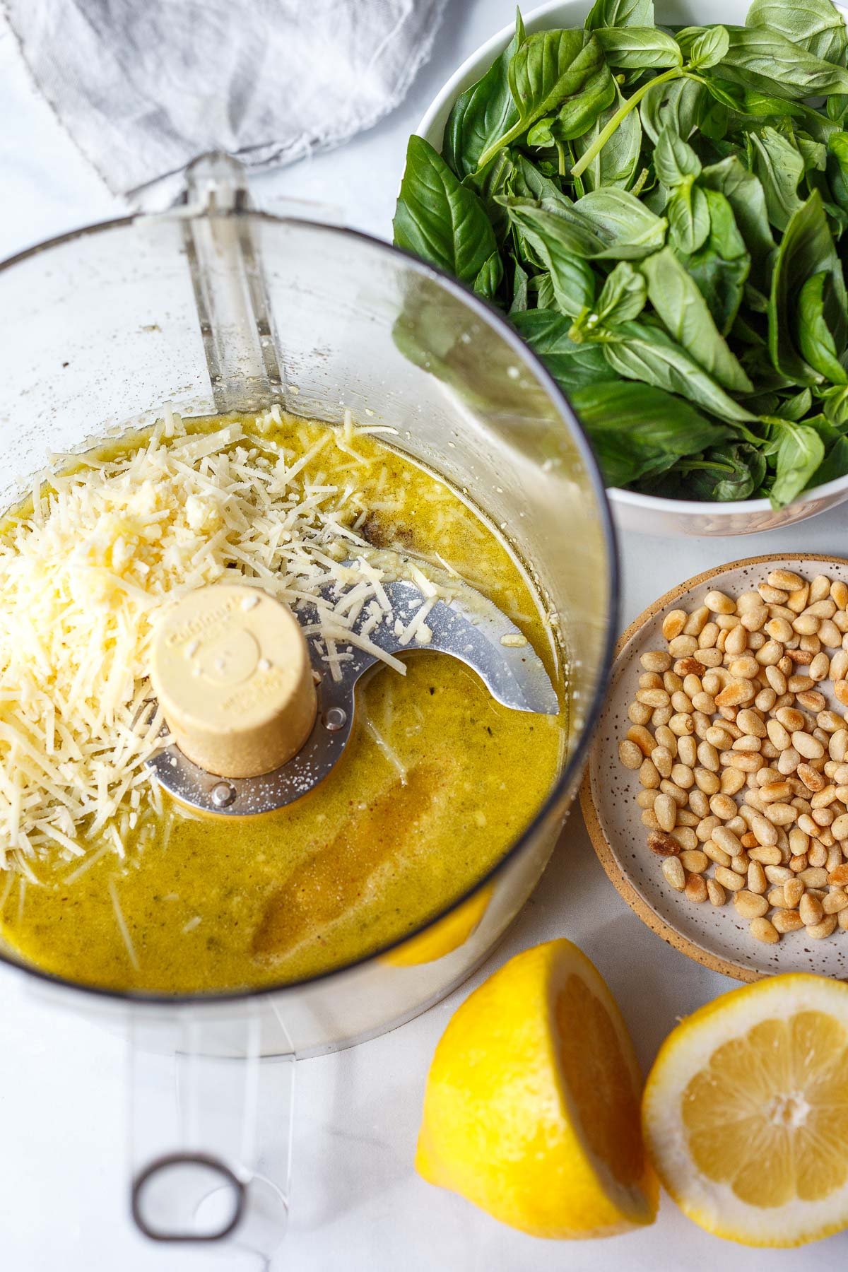 food processor with olive oil, lemon juice, and parmesan next to bowl of basil and plate of pine nuts. 