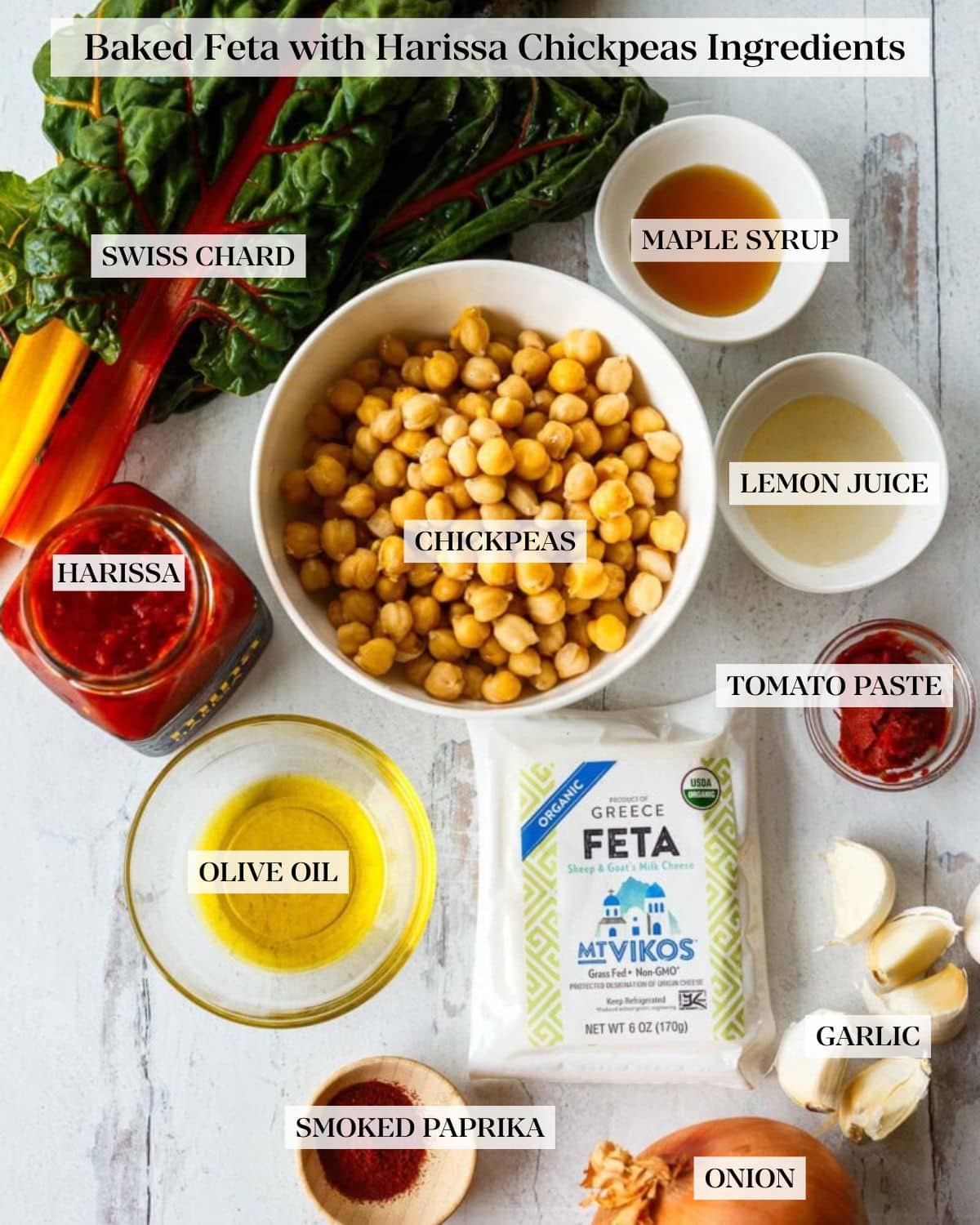  baked feta with harissa chickpeas ingredients: labeled and arranged on a white wood surface - swiss chard, maple syrup, lemon juice, chickpeas, harissa, tomato paste, olive oil, feta, garlic, smoked paprika, and onion.