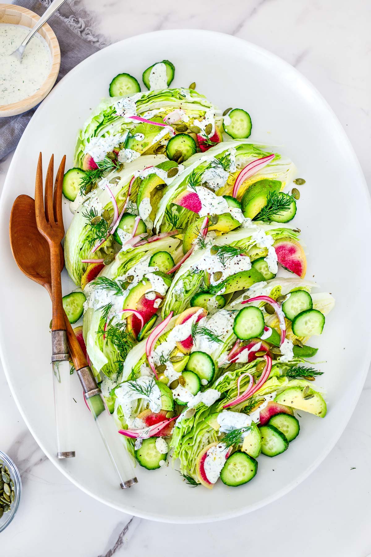 oval platter with wedge salad with red onion, cucumber, avocado, pepitas, watermelon radish, and fresh dill, topped with creamy dressing, served with wood utensils.
